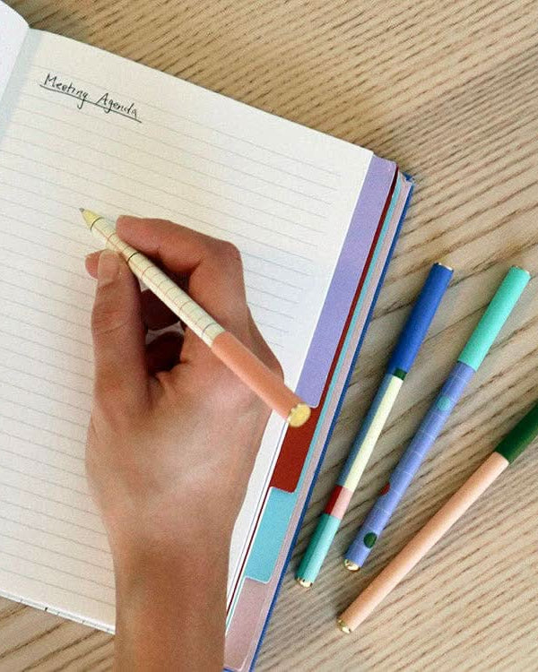 Four pens and a notebook opened and someone using one of the pens to write in notebook