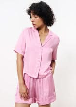 Woman wearing pink short sleeve cropped satin blouse with boxy shape with collar revere, v neck and ivory buttons.