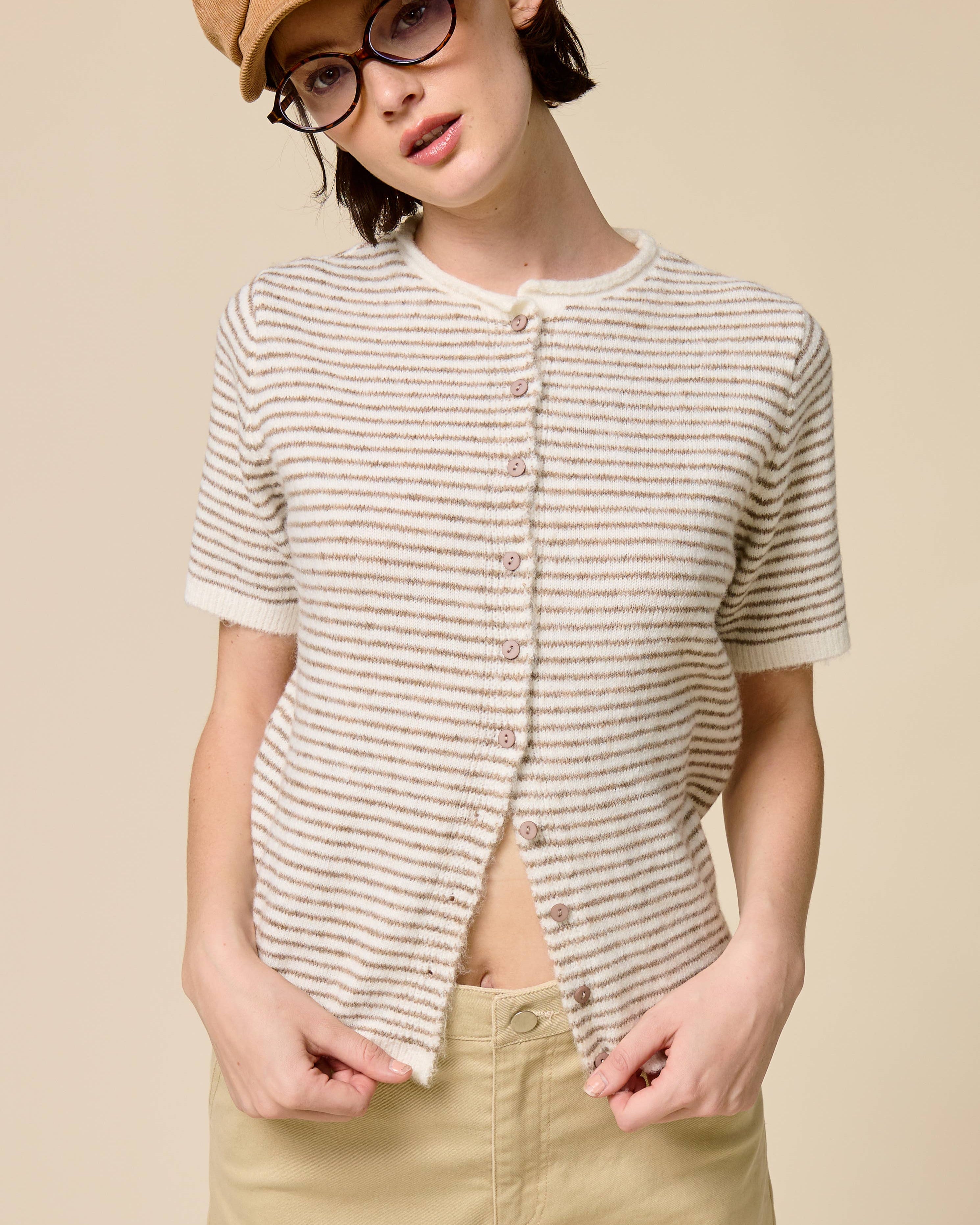 Person wearing a striped shirt, beige cap, and glasses on a beige background