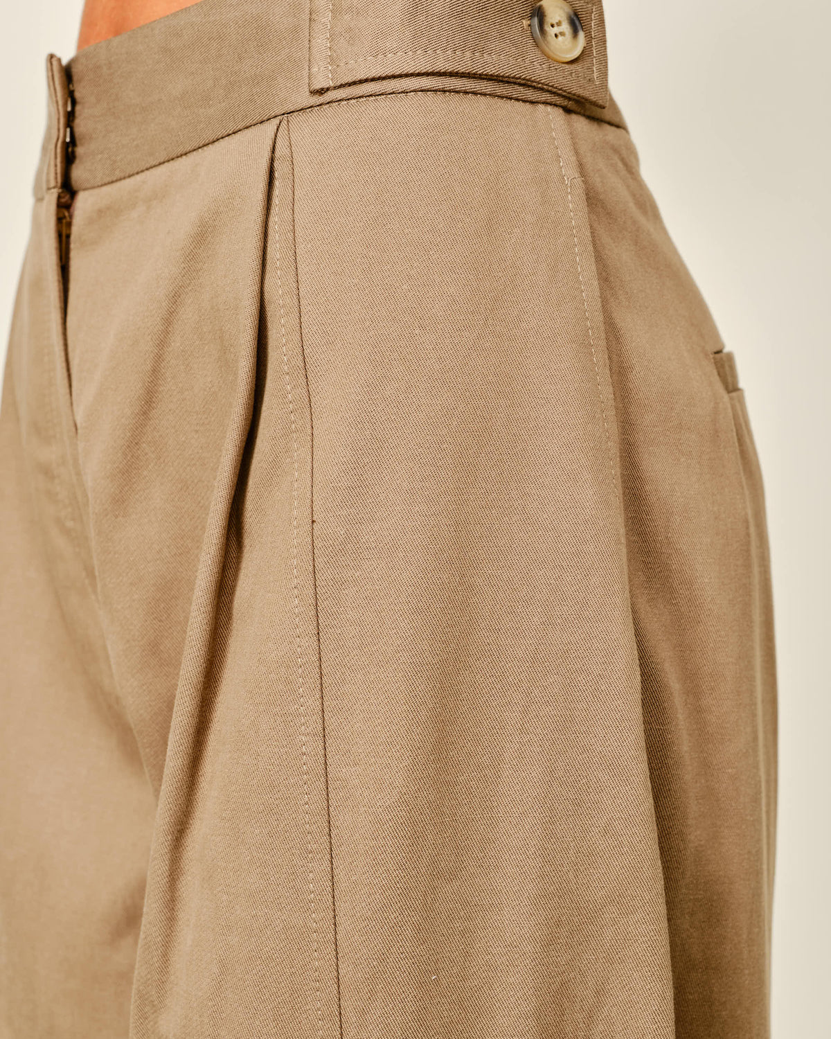 Close-up of a model wearing high-waisted taupe trousers, showing the waistband with a button closure and pleated front detail.