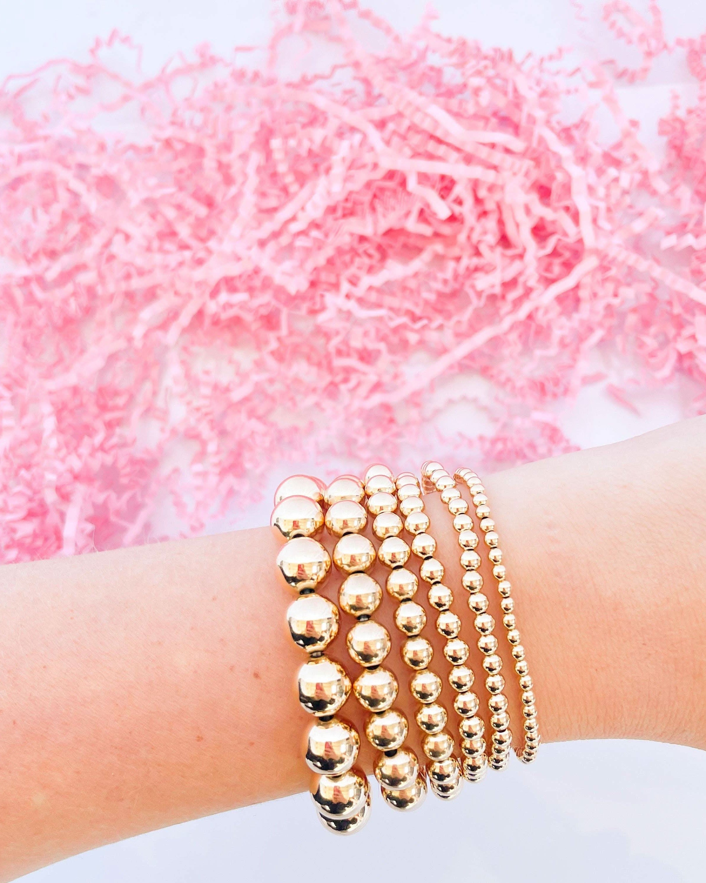 Gold beaded bracelets on a wrist with a pink textured background