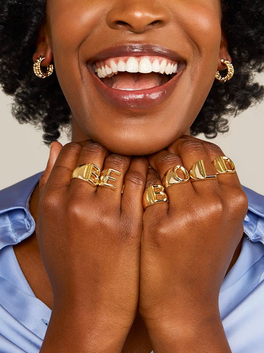 Smiling woman wearing blue shirt with hands under her chin wearing gold letter rings in the words BE  BOLD
