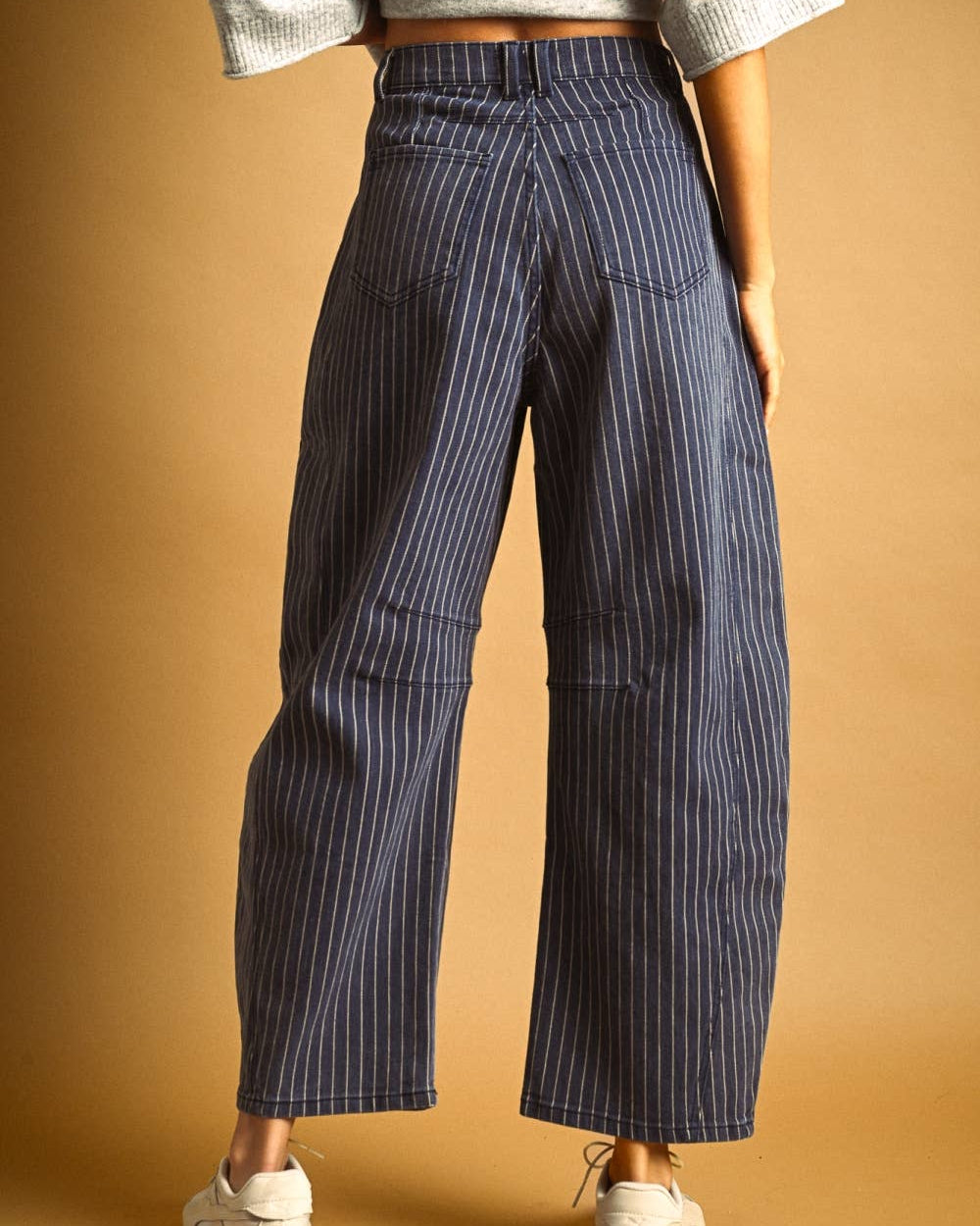 A person shown from the back wearing high-waisted, wide-leg cropped pants with thin vertical pinstripes, paired with a light gray knit top and white sneakers, against a warm tan studio backdrop.