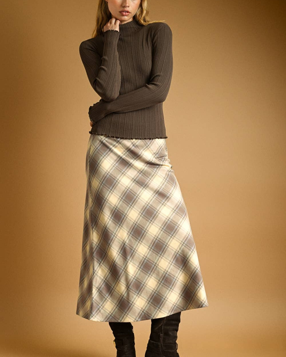 Woman wearing a brown ribbed sweater and plaid skirt against a beige background