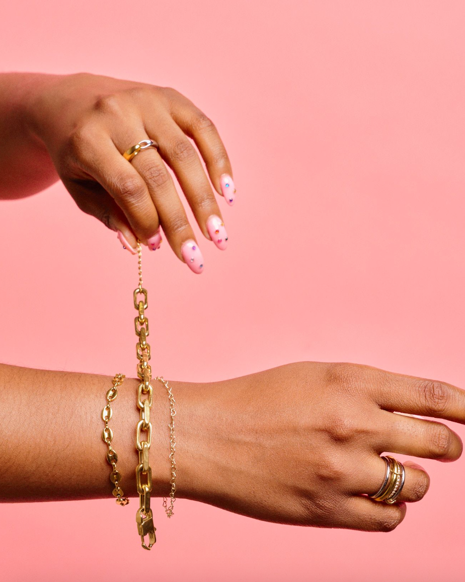 Woman putting on a Gold, waterproof, chunky link bracelet from Nikki smith against a pink surface