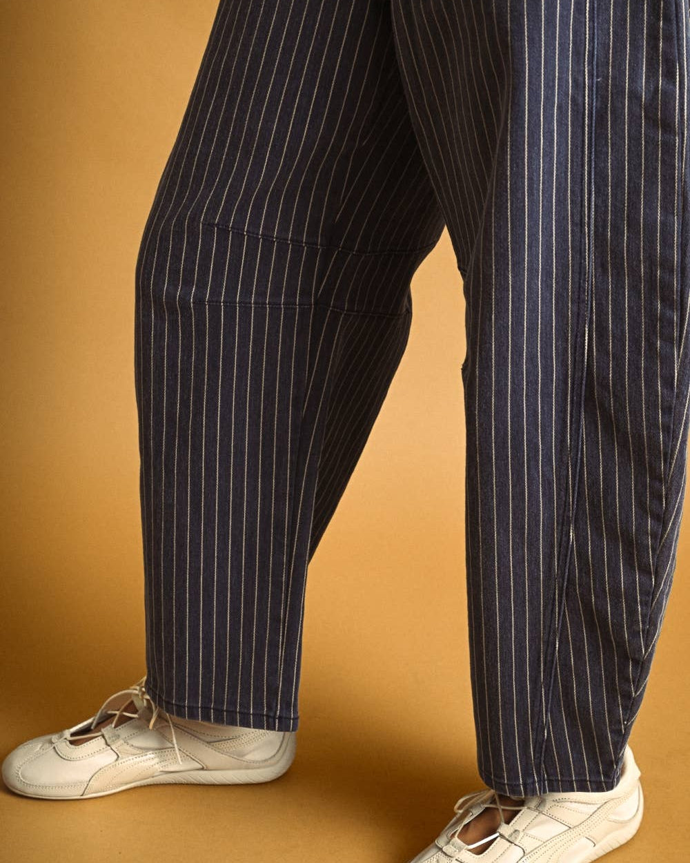 A person wearing loose navy blue pinstriped trousers and white sneakers, standing against a warm tan studio background.