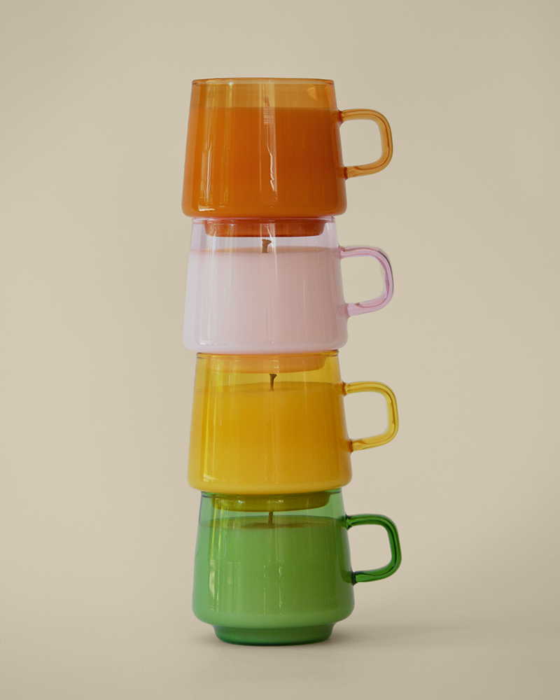 Four candles in orange, pink, yellow, and green opaque reusable mug sitting stacked on one another in the sun against a cream colored wall