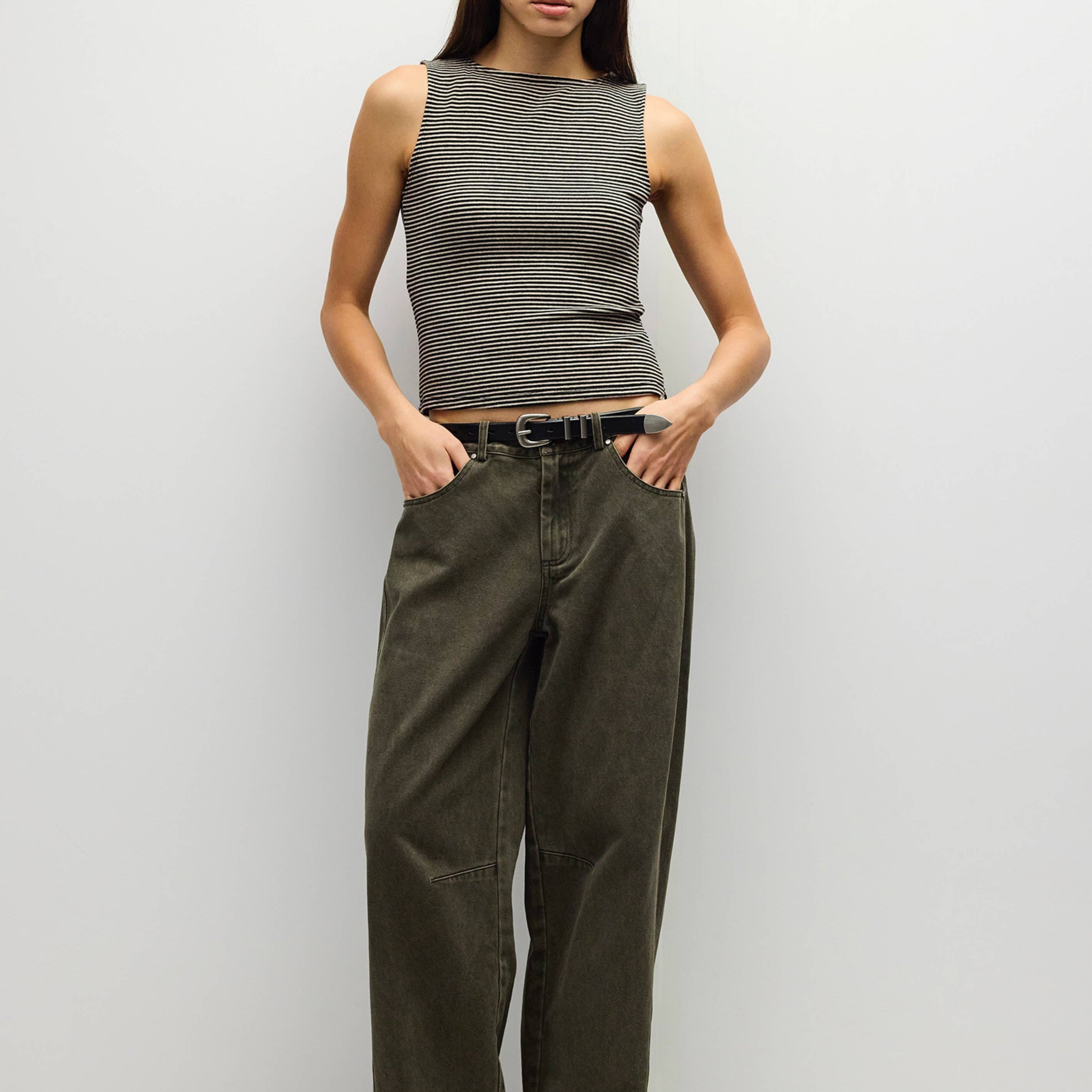 A woman stands against a plain light gray studio wall wearing a sleeveless ribbed gray top, dark olive wide-leg pants, and white shoes.