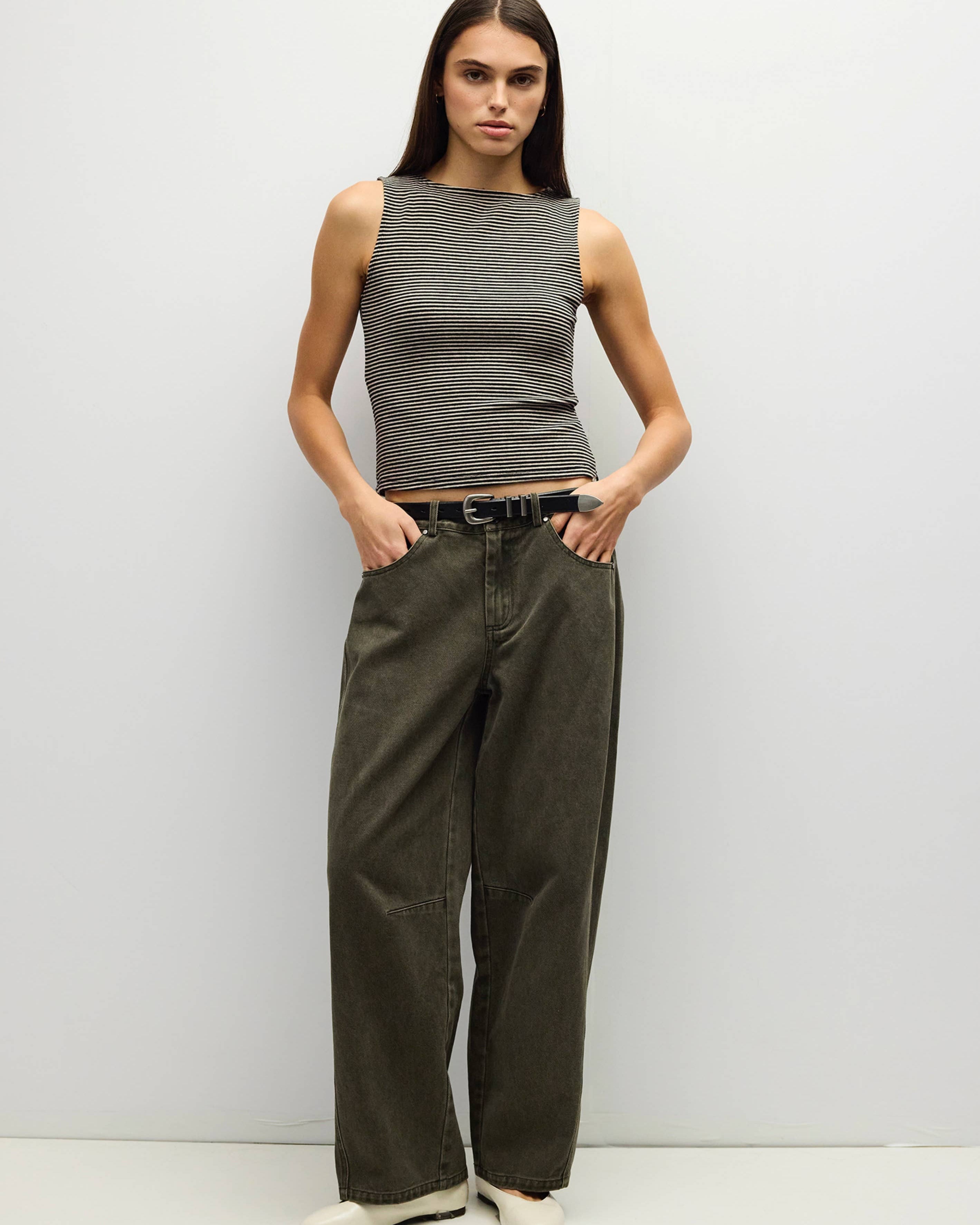 A woman stands against a plain light gray studio wall wearing a sleeveless ribbed gray top, dark olive wide-leg pants, and white shoes.