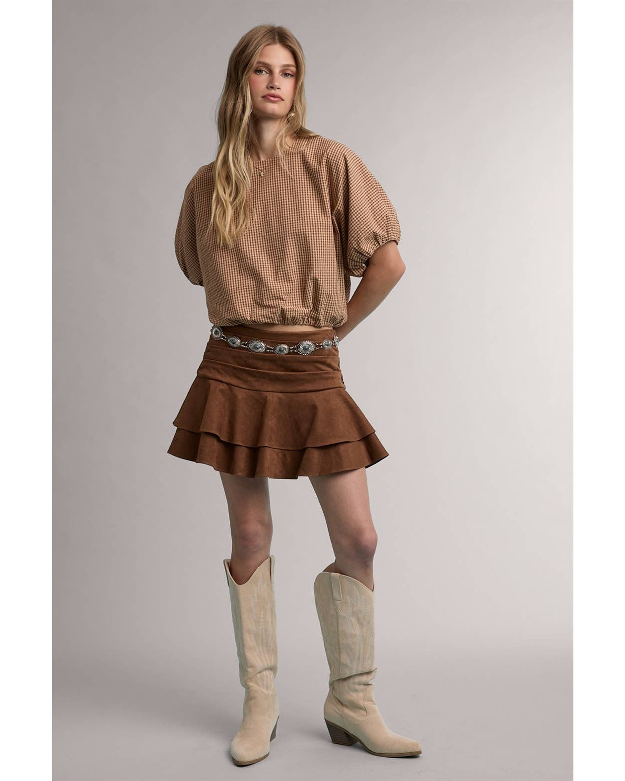 A woman modeling a brown textured short-sleeve blouse with puffed sleeves, a brown tiered ruffle mini skirt with a silver-toned patterned belt, and beige cowboy boots, set against a plain light gray studio background.