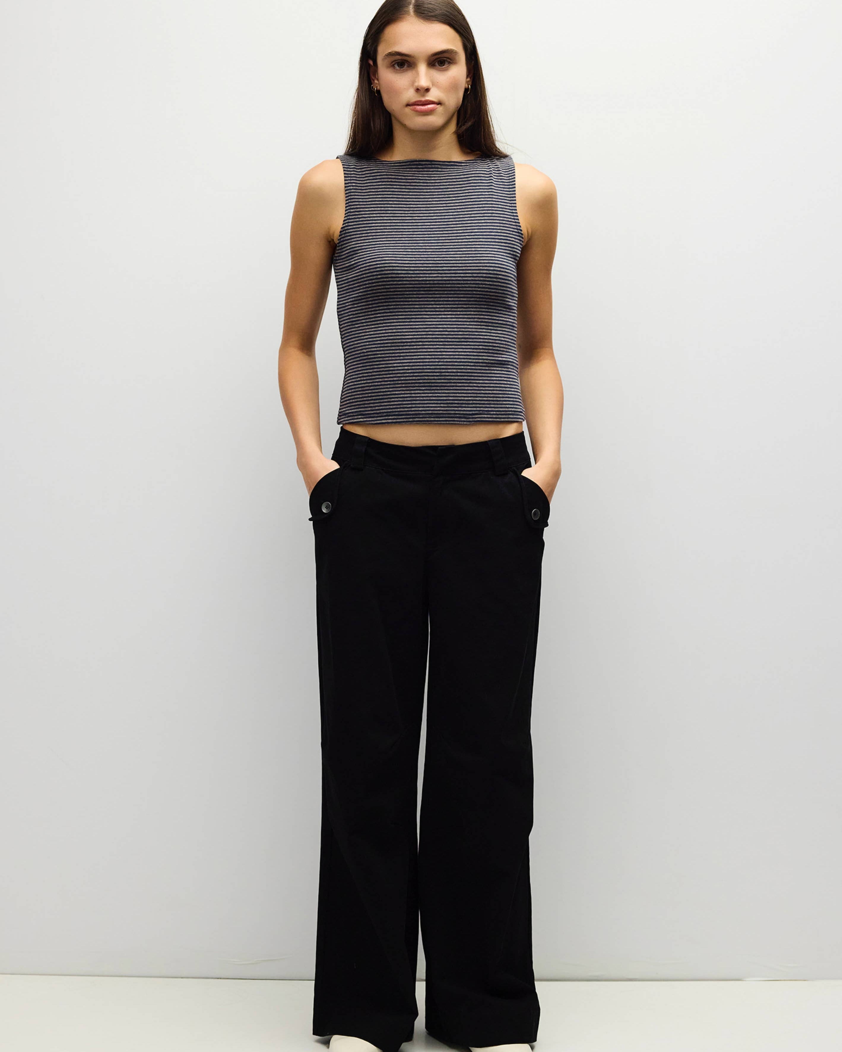 A woman stands against a plain light gray studio background wearing a sleeveless dark gray ribbed knit top and black wide-leg pants with white shoes.