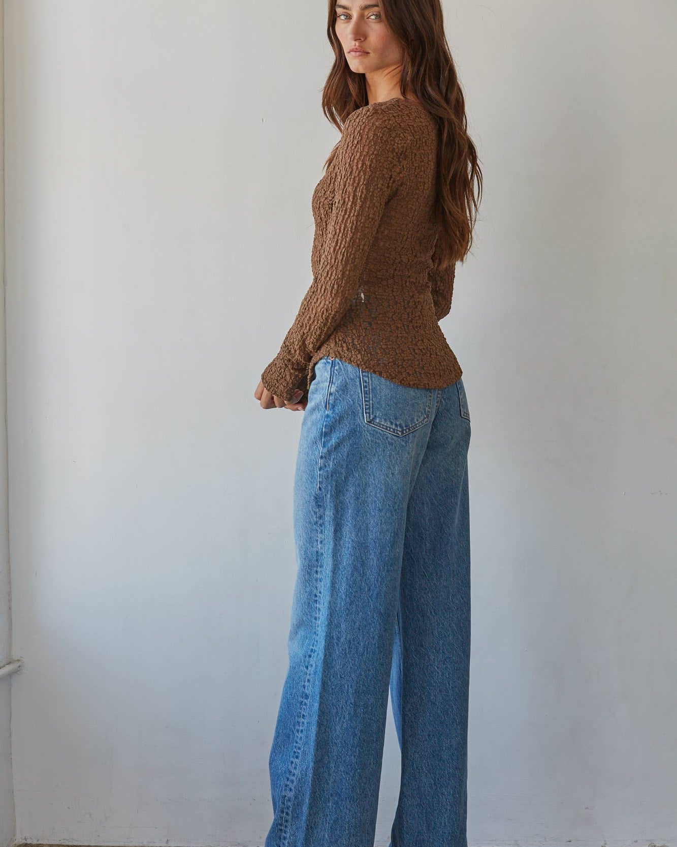 A woman stands in profile wearing a brown textured knit sweater and wide-leg blue jeans against a plain off-white wall.