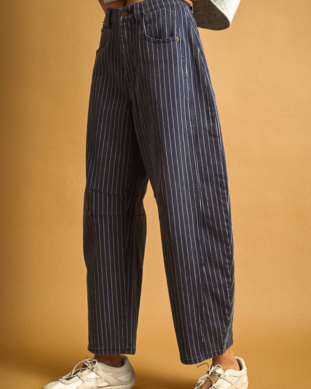 A person wearing navy blue pinstriped wide-leg jeans, a light gray knit top, and white sneakers, standing against a warm tan studio background.