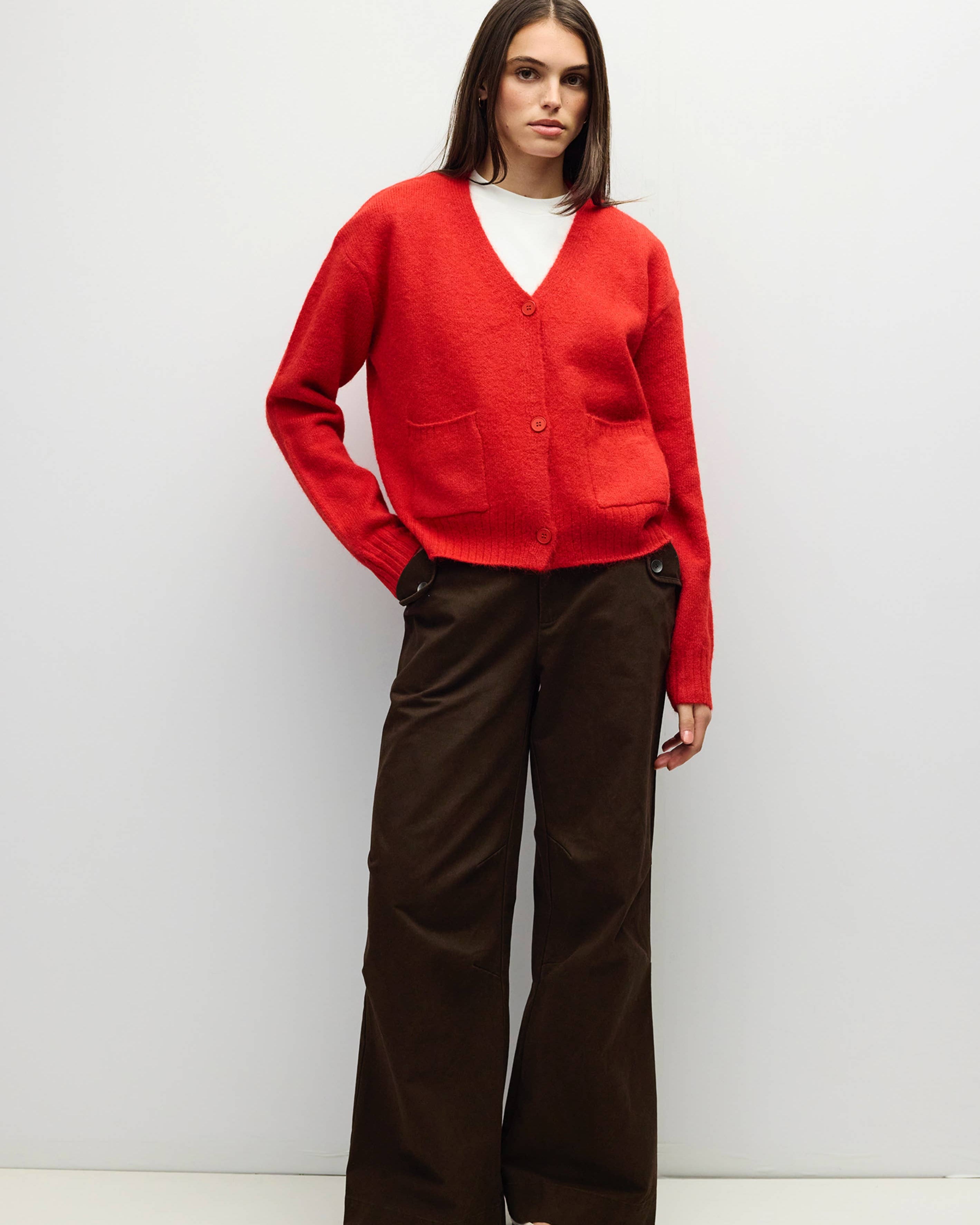 A woman stands against a light gray studio background wearing a bright red knit cardigan over a white top, paired with dark brown wide-leg trousers and white pointed shoes.