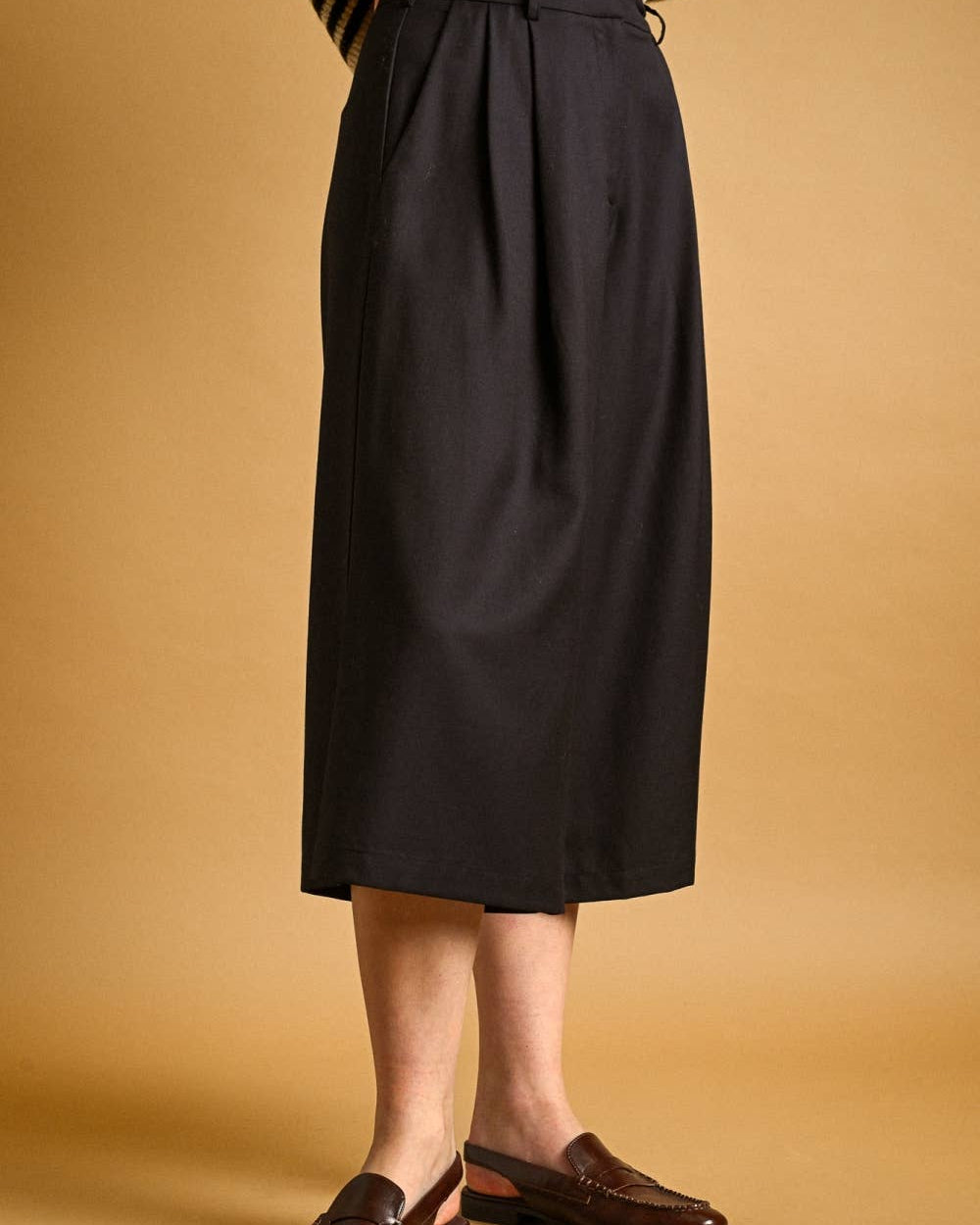 A person wearing a dark charcoal pleated midi skirt, a black and white striped sweater, and brown leather loafers, standing against a solid tan background.