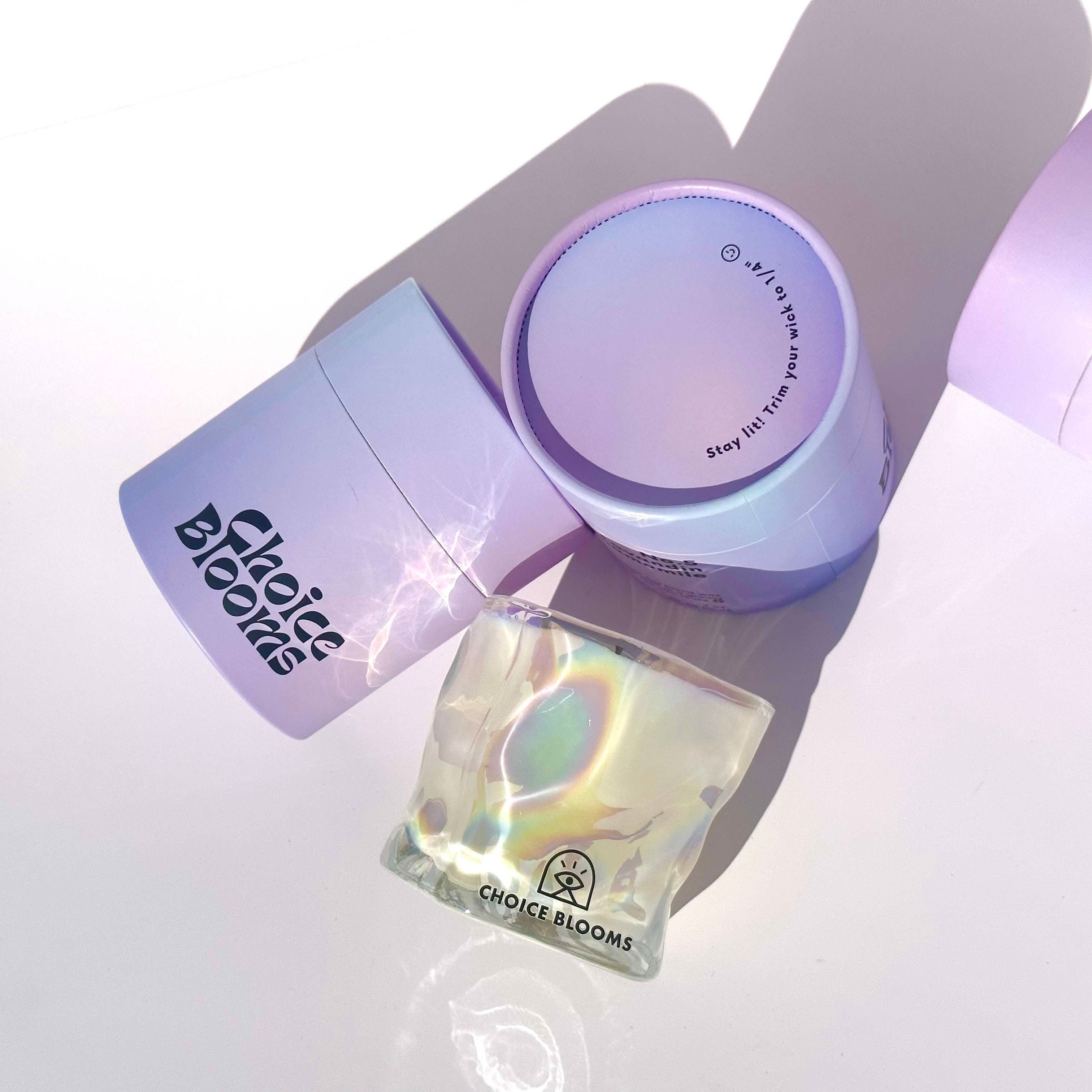 One candle in iridescent wavy glass cup laying on its side next to two purple packaging containers