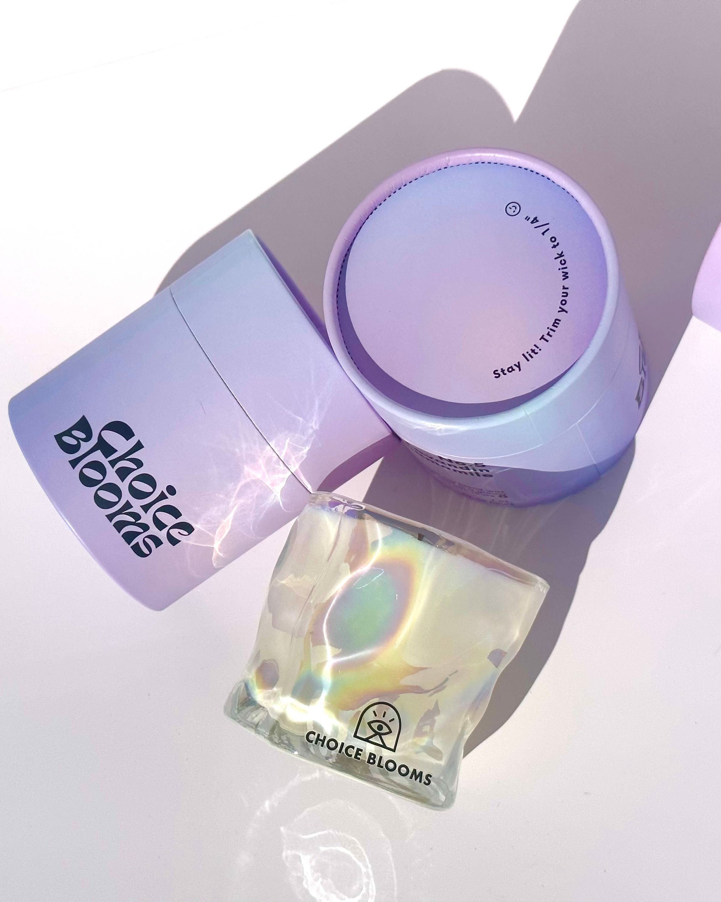 One candle in iridescent wavy glass cup laying on its side next to two purple packaging containers