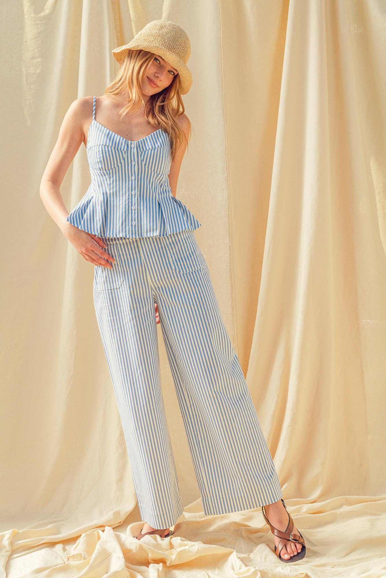 Woman wearing a blue striped top and white pants with a straw hat against a beige curtain background