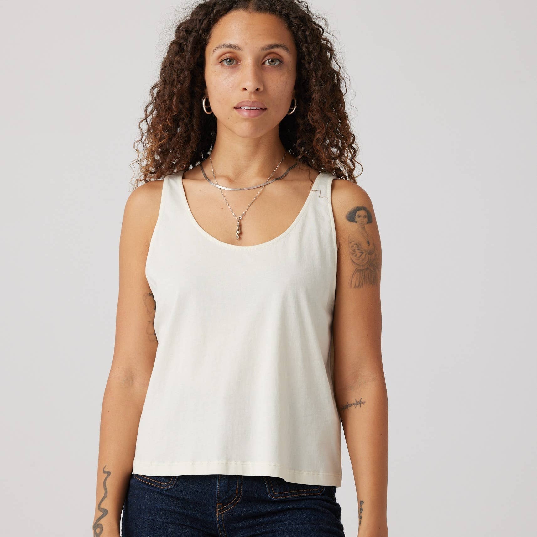 Woman wearing a white tank top and dark jeans against a plain background
