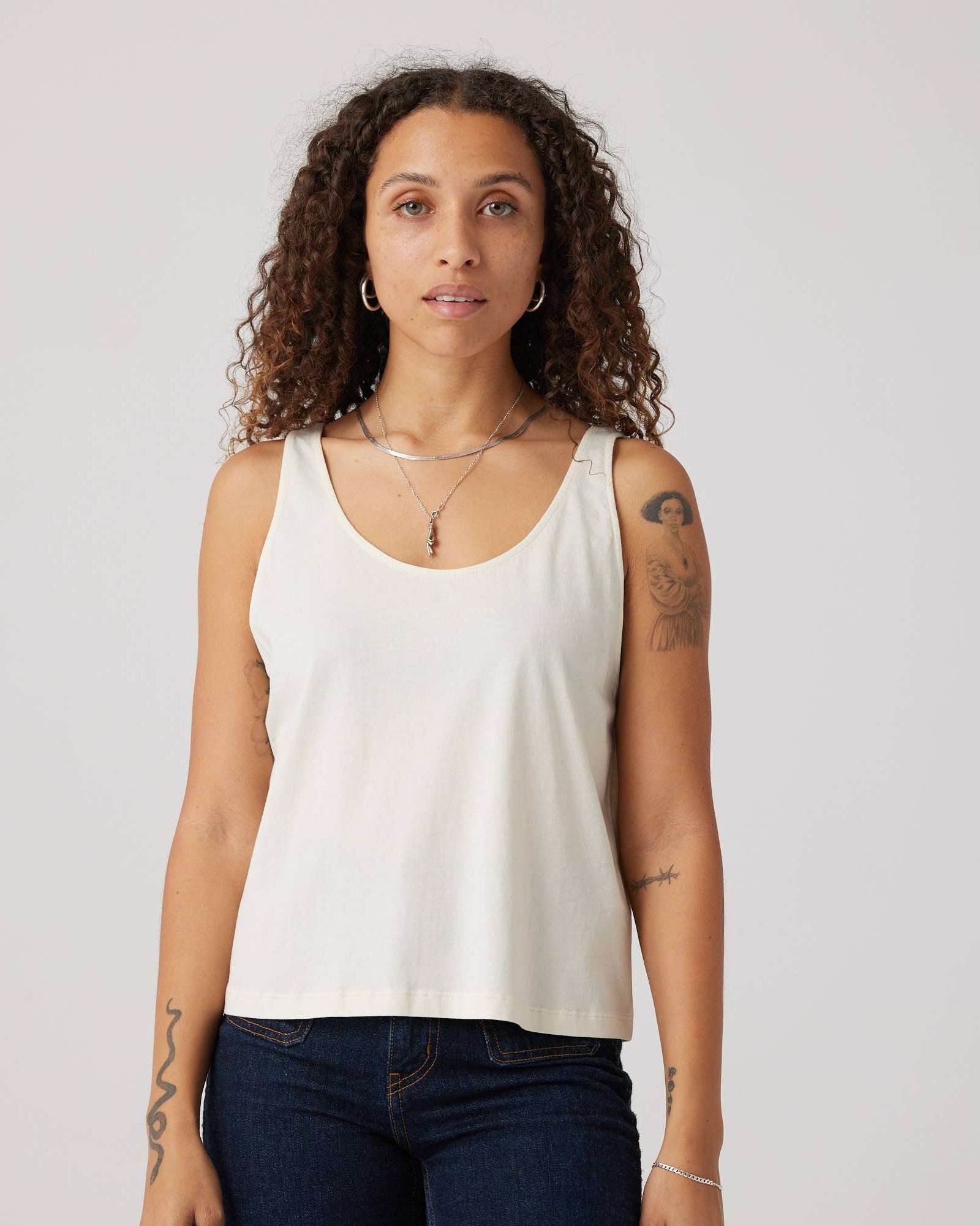Woman wearing a white tank top and dark jeans against a plain background