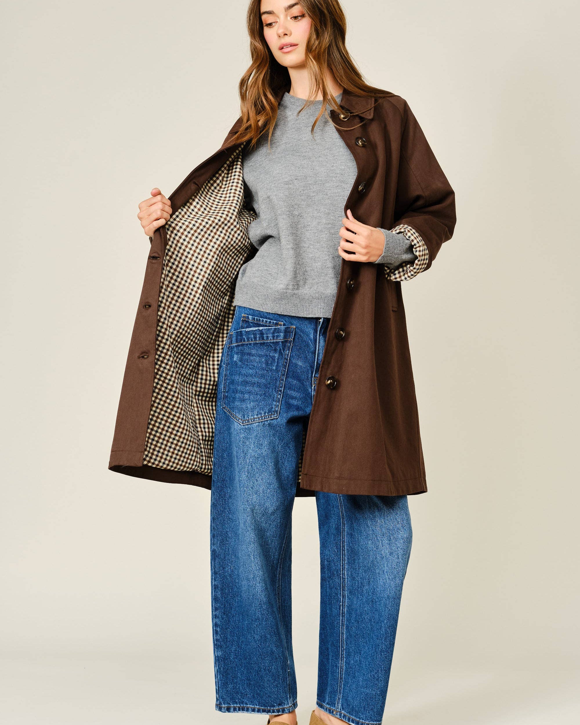 Woman wearing a brown coat over a gray sweater and blue jeans on a white background