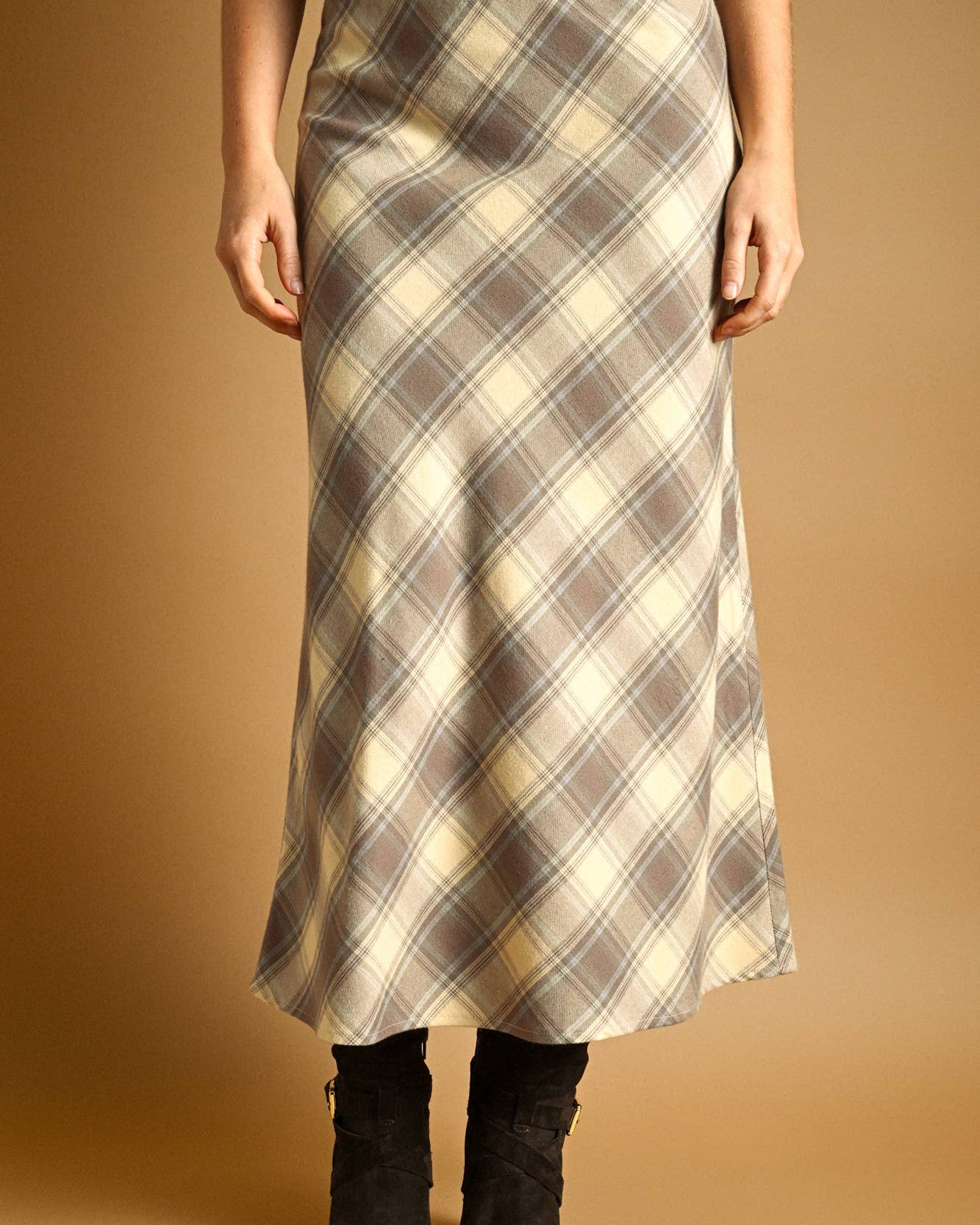 A woman wearing a long plaid midi skirt in beige, brown, and cream tones, paired with black ankle boots, against a solid warm brown studio background.