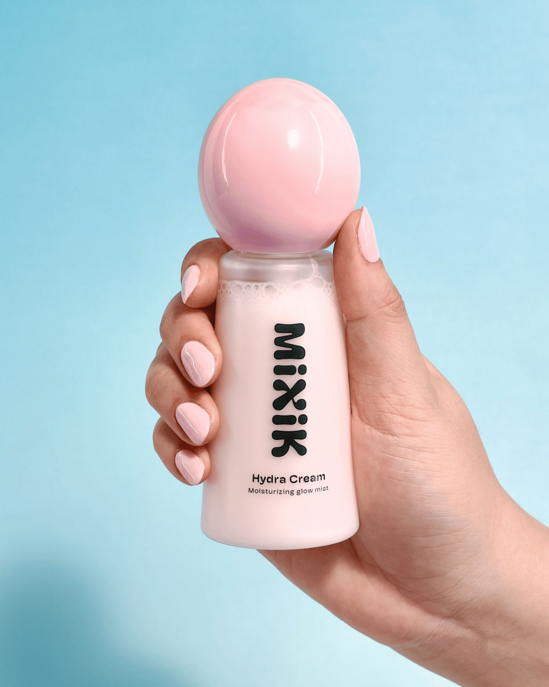 Hand holding a pink bottle of Mixik Hydra Cream against a light blue background