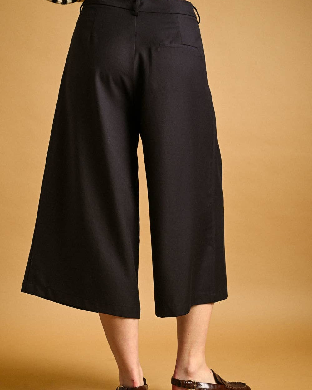 A person wearing black wide-leg cropped pants with a striped top and brown sandals, standing against a warm tan studio background.