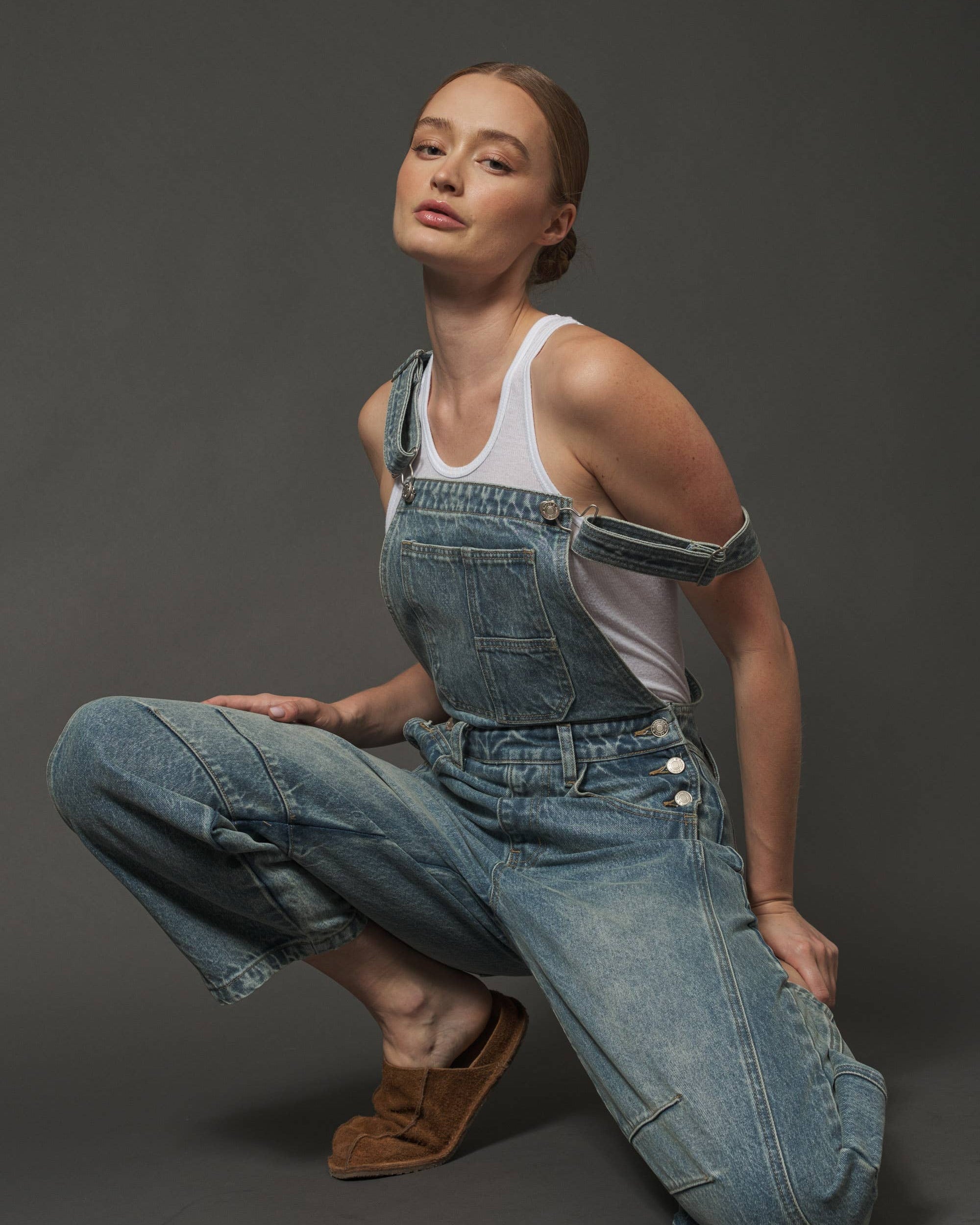 Person wearing blue denim overalls and a white tank top on a dark background