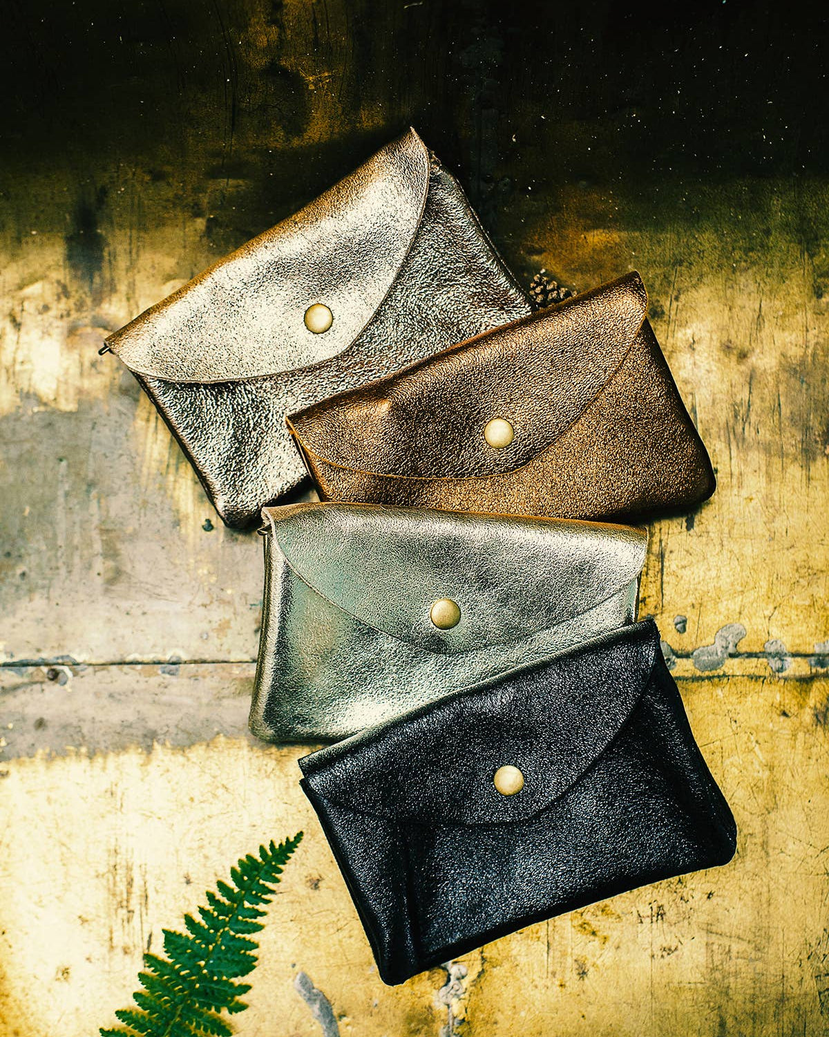 Four small leather envelope-style pouches or card cases in metallic silver, bronze, gray, and black, arranged diagonally on a distressed gold surface with a green fern leaf at the lower left.