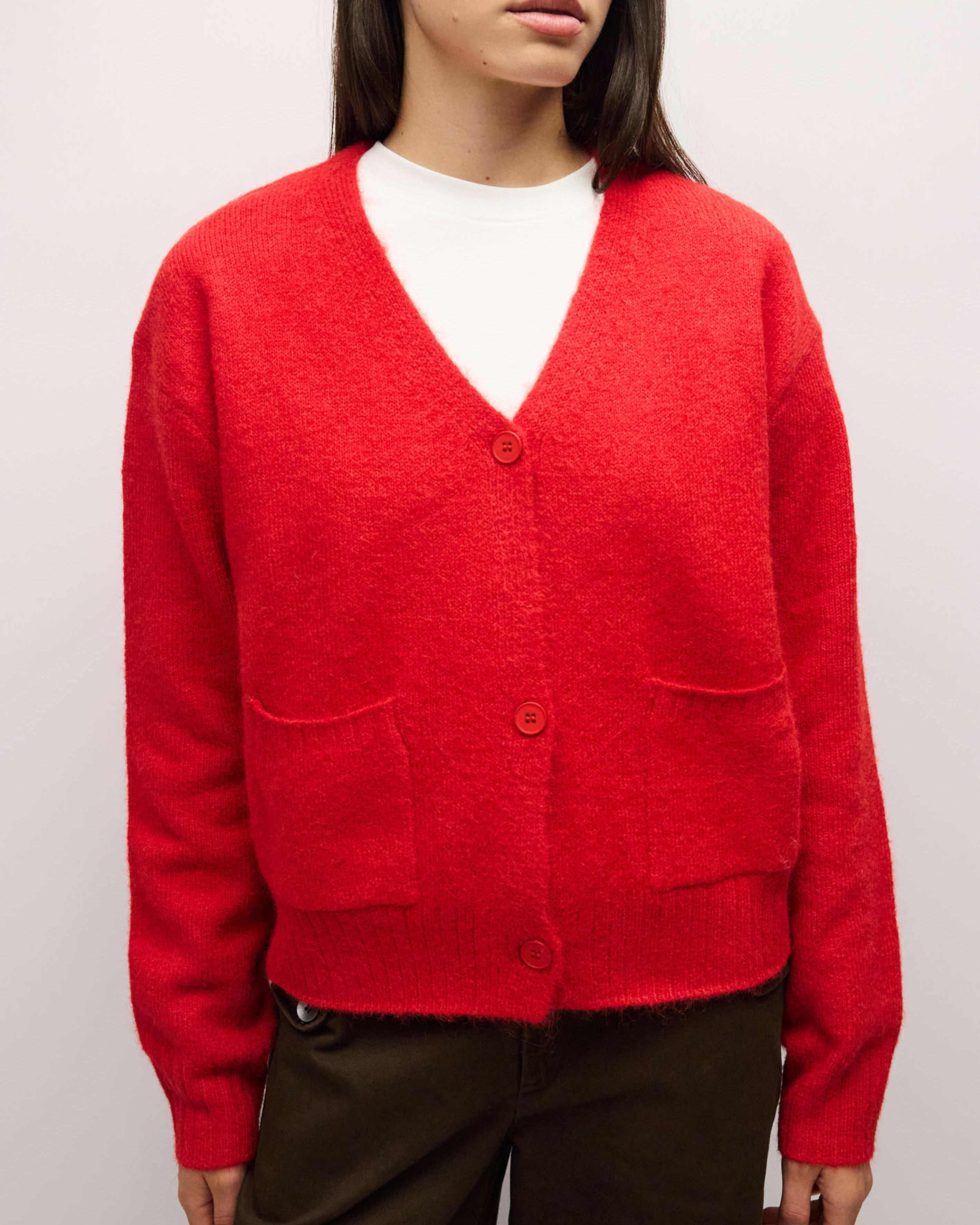 A woman wearing a bright red fuzzy knit cardigan with a V-neck, three buttons, and front patch pockets, layered over a white top and dark brown pants, photographed against a light gray studio background.