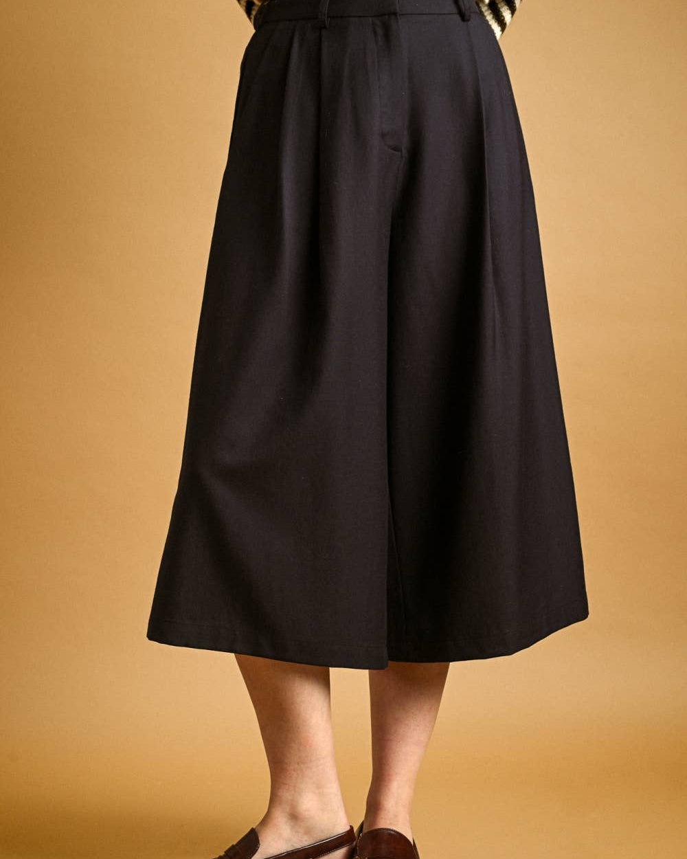 A person wearing a black wide-leg midi skirt, styled with a black and white striped top and brown leather loafers, against a solid warm tan background.