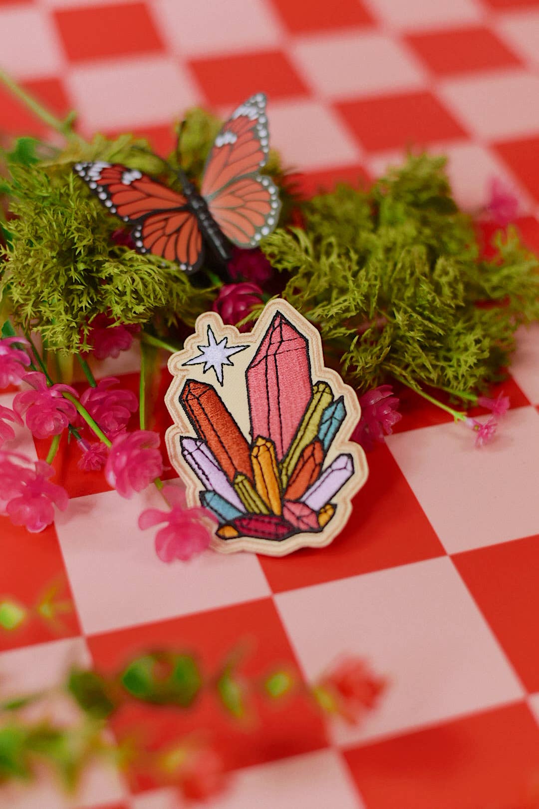 A colorful enamel pin shaped like a cluster of crystals with a butterfly decoration, displayed on a red and white checkered surface with green moss and pink flowers.