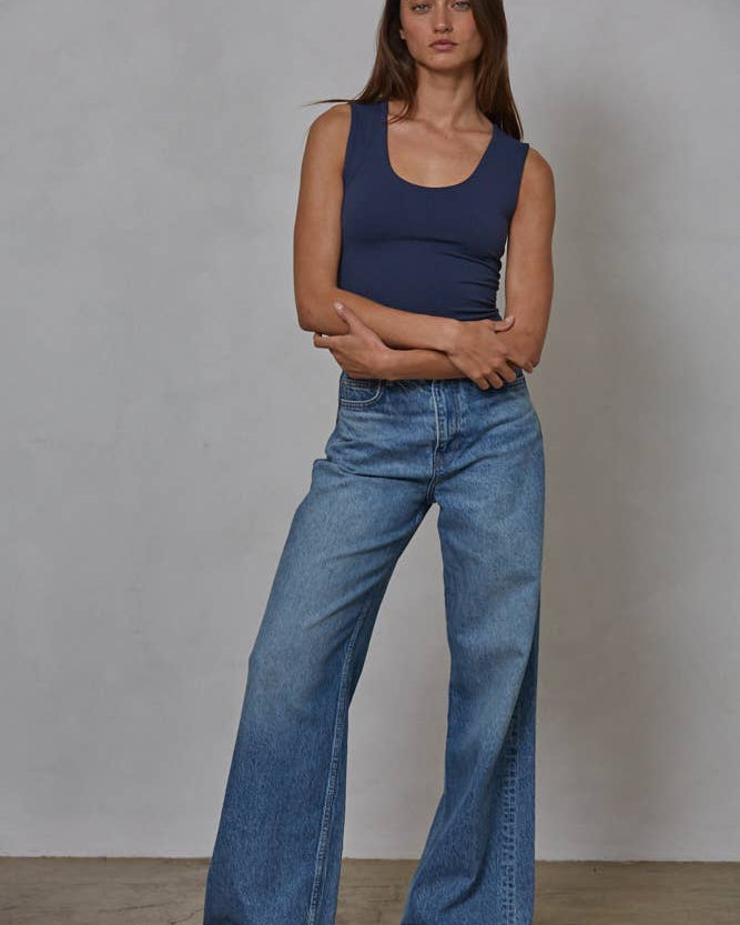 Woman wearing a blue tank top and jeans standing against a plain background