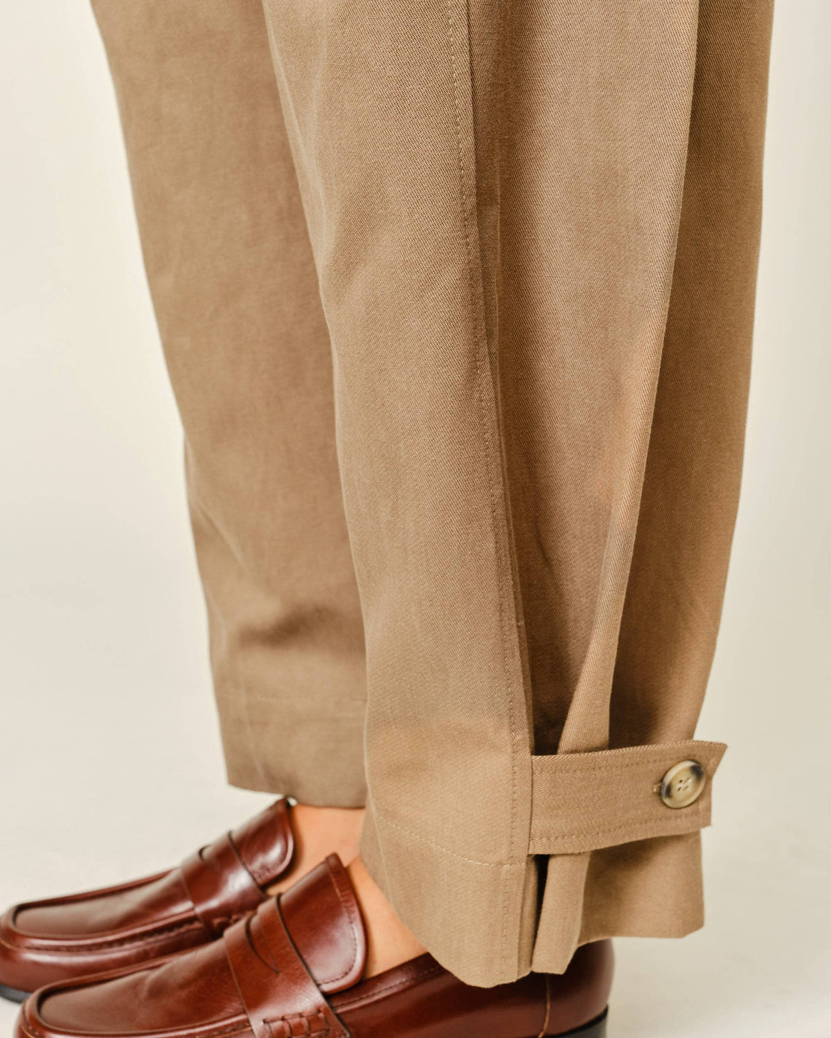 Close-up of a person wearing tan trousers with a buttoned cuff and brown leather penny loafers, shown against a plain light beige background.
