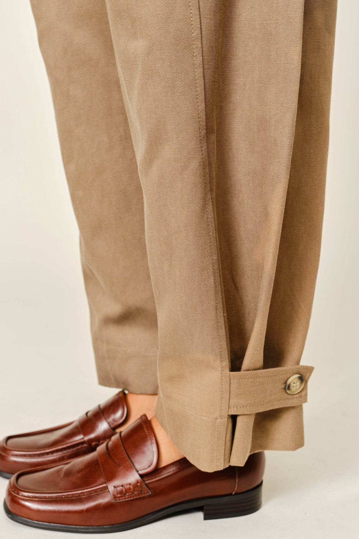 Close-up of a person wearing tan trousers with a buttoned cuff and brown leather penny loafers, shown against a plain light beige background.