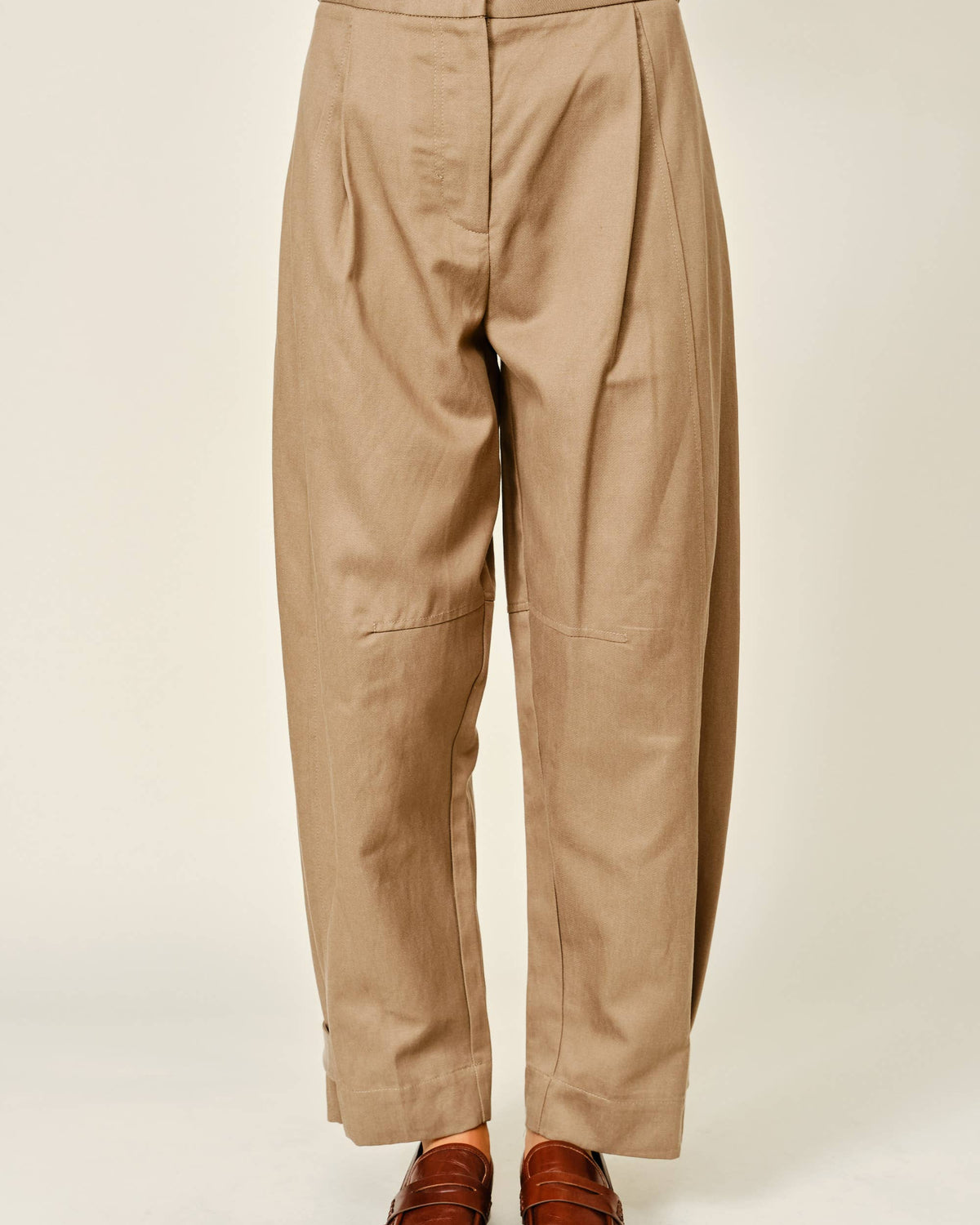 A person shown from the waist down wearing loose, high-waisted tan pants with front pleats and brown leather loafers, photographed against a plain light beige studio background.