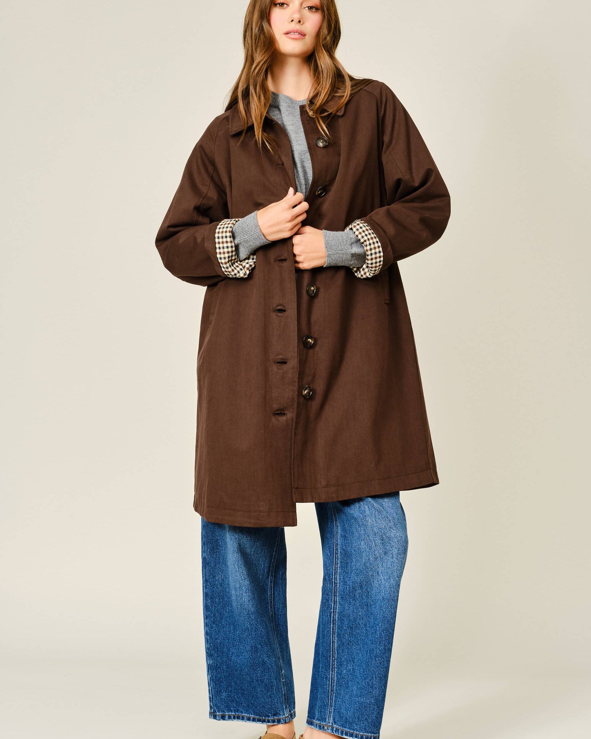 Woman wearing a brown coat, gray sweater, blue jeans, and brown shoes on a beige background