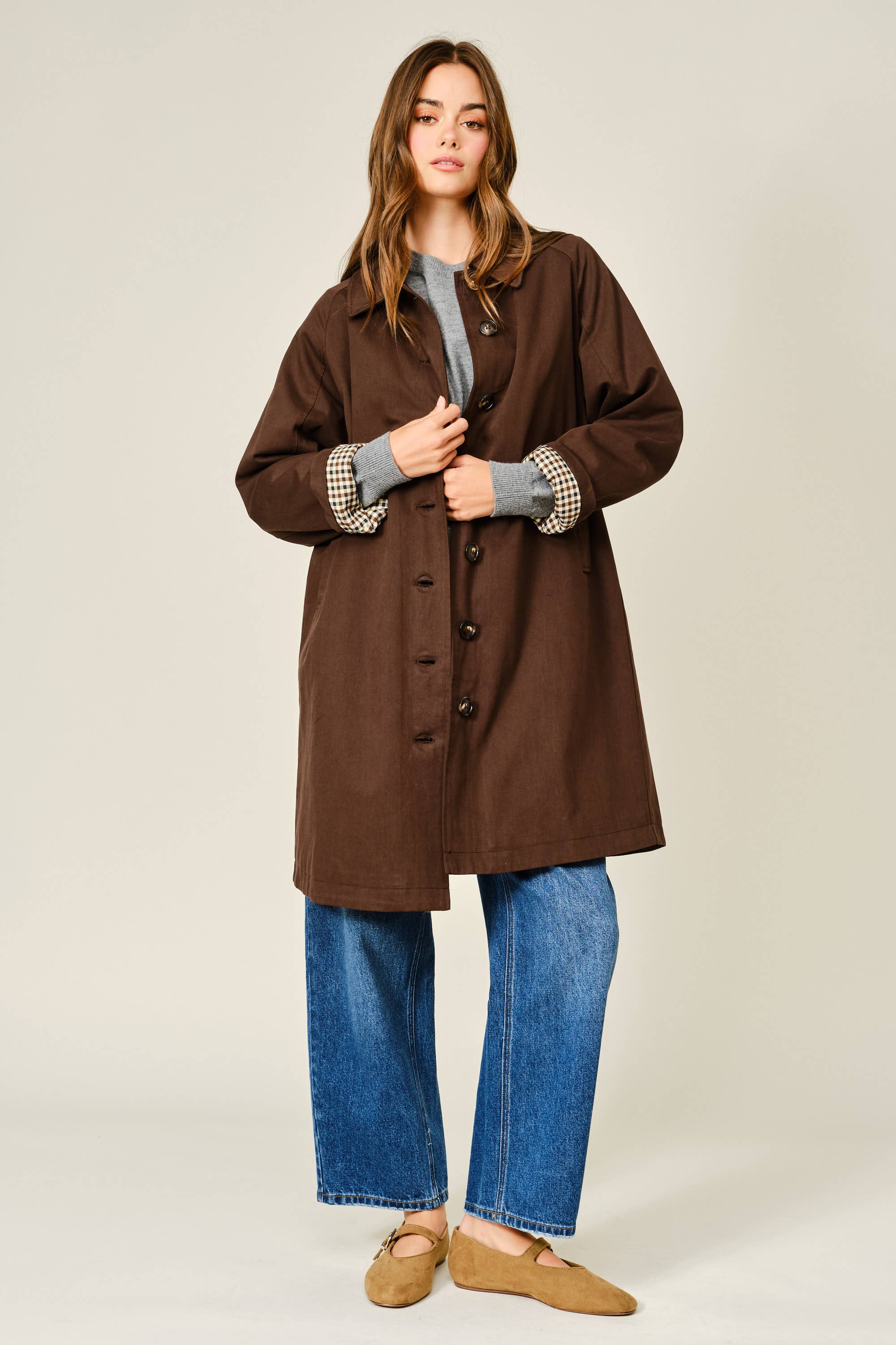 Woman wearing a brown coat, gray sweater, blue jeans, and brown shoes on a beige background