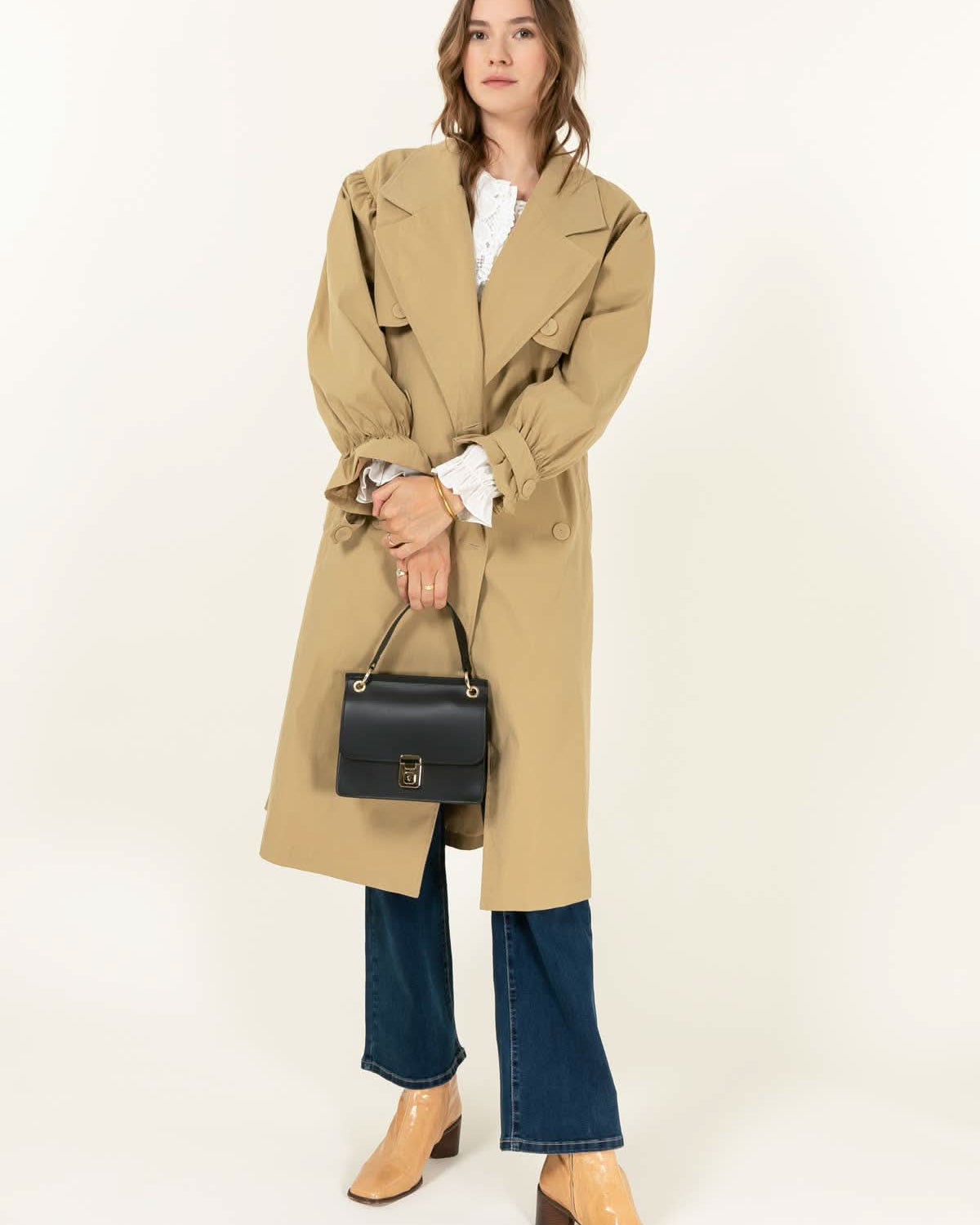 Woman wearing brown trench coat and holding Black leather handbag with gold details and clasp sitting against white background