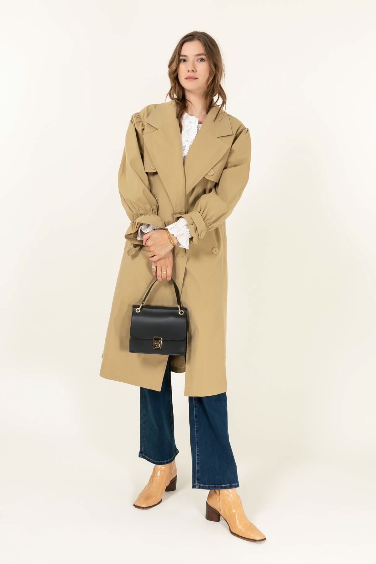 Woman wearing brown trench coat and holding Black leather handbag with gold details and clasp sitting against white background