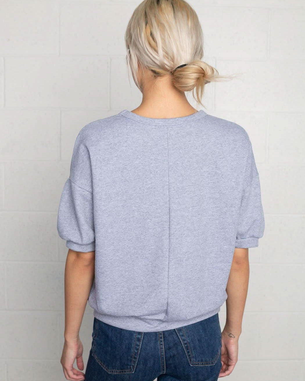 Woman facing away from the camera wearing natural grey, short sleeve, puff sleeve, blouse t-shirt top, with vertical indent line down the back