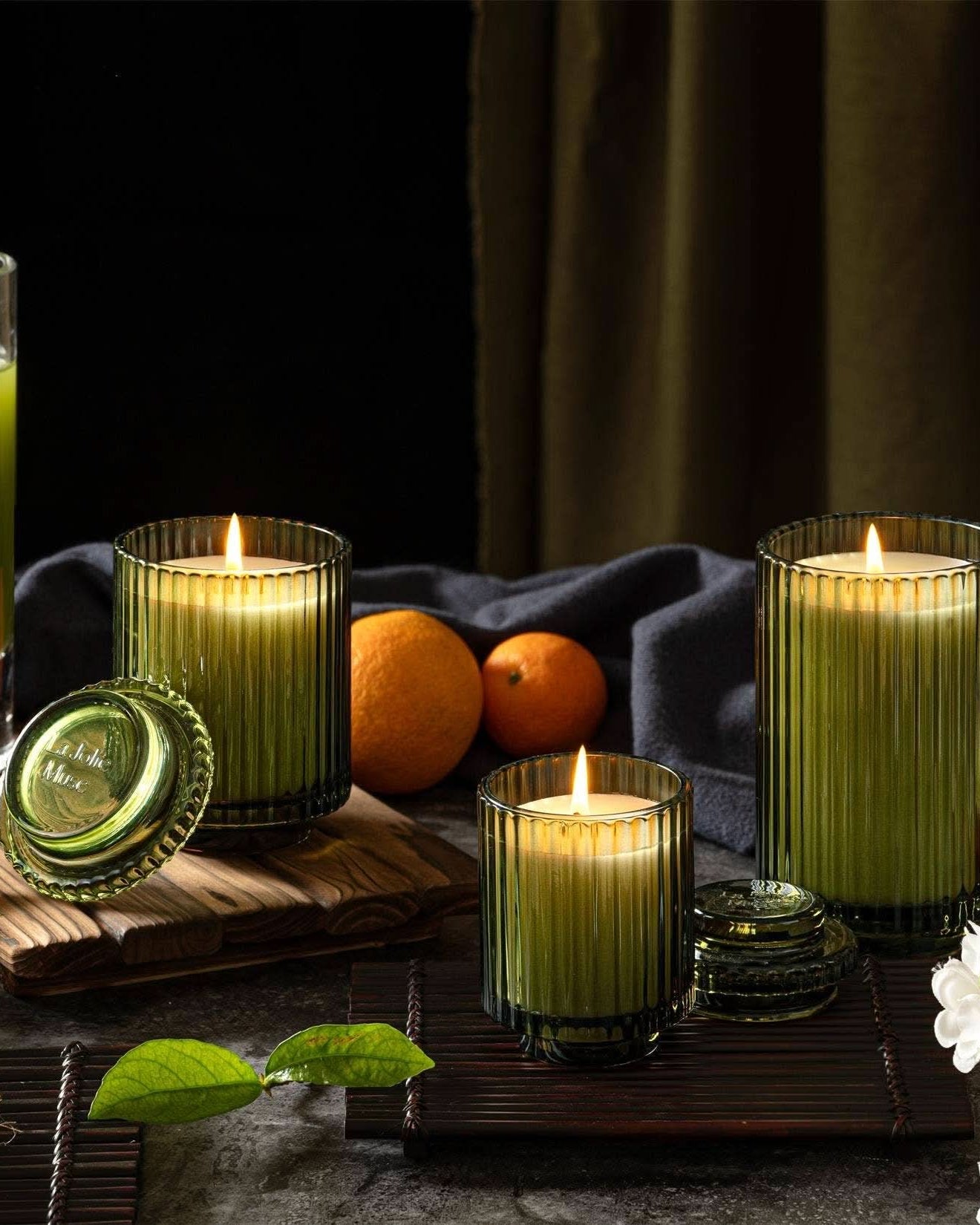 Green candles in glass holders with a glass of green juice and oranges on a dark background