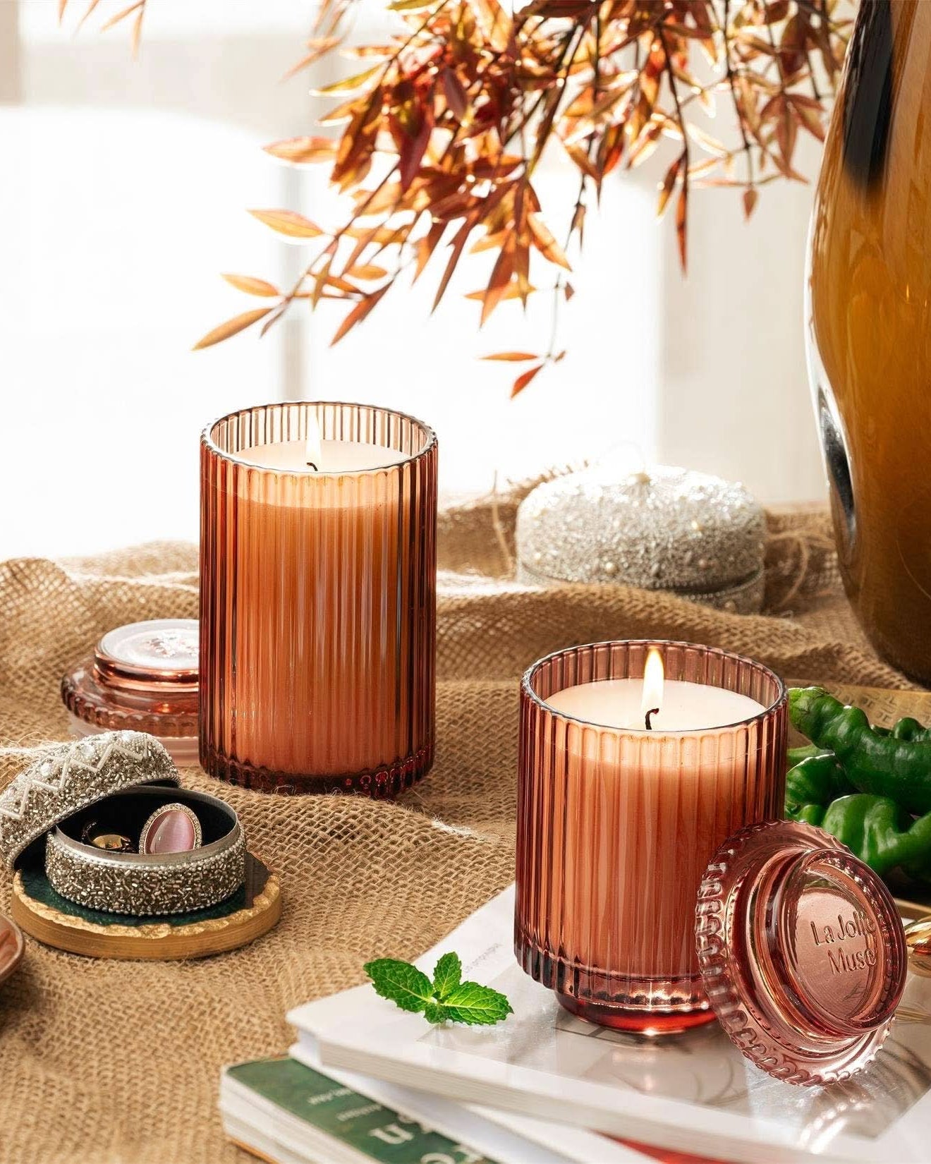 Decorative candles in brown glass containers on a table with books and a vase.