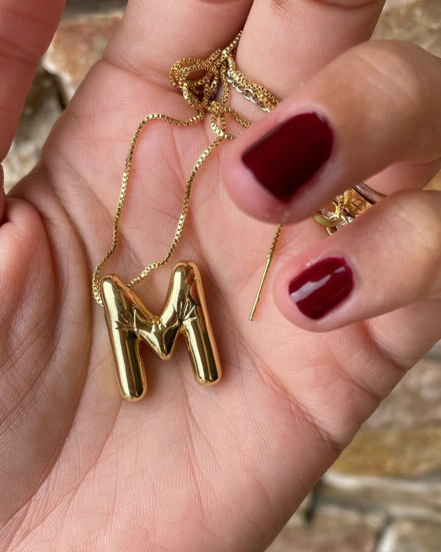 Gold necklace with a 'M' pendant held in a hand with red nail polish.