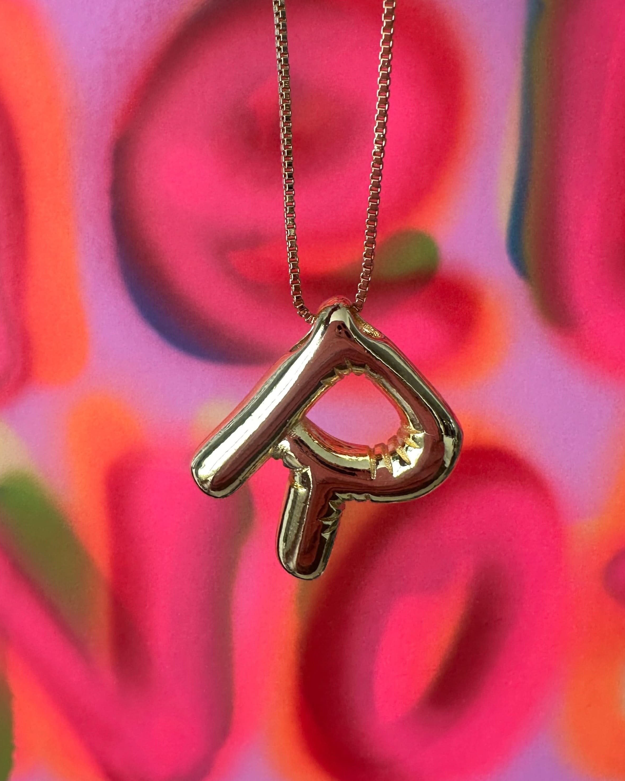 Gold necklace with a heart-shaped R pendant against a colorful