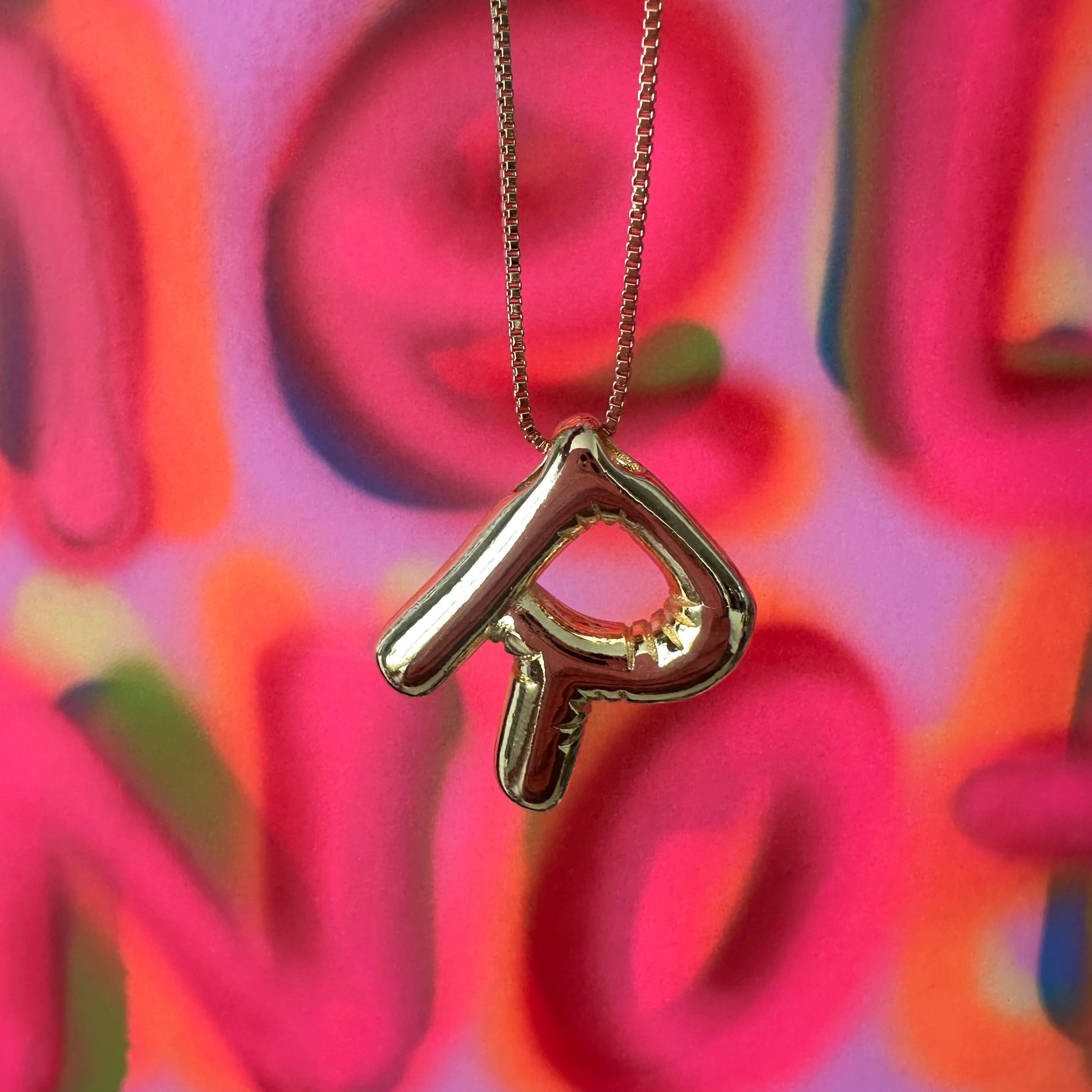 Gold necklace with a heart-shaped R pendant against a colorful