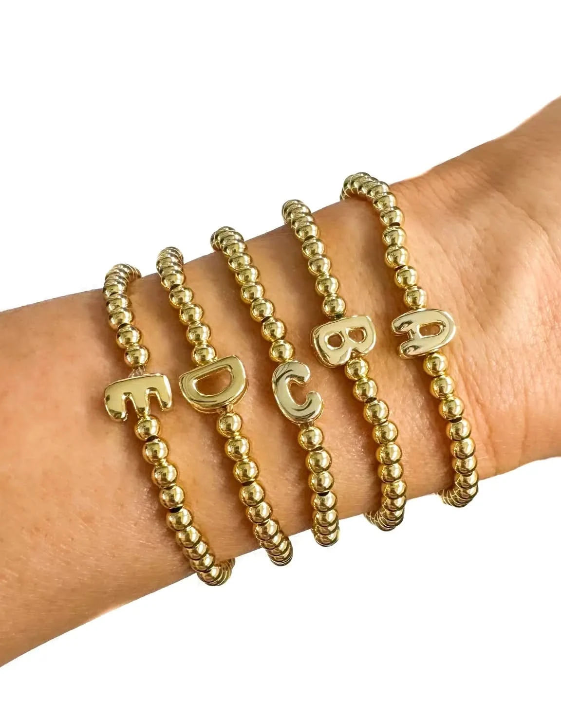 Gold beaded bracelets with letter charms on a wrist against a white background