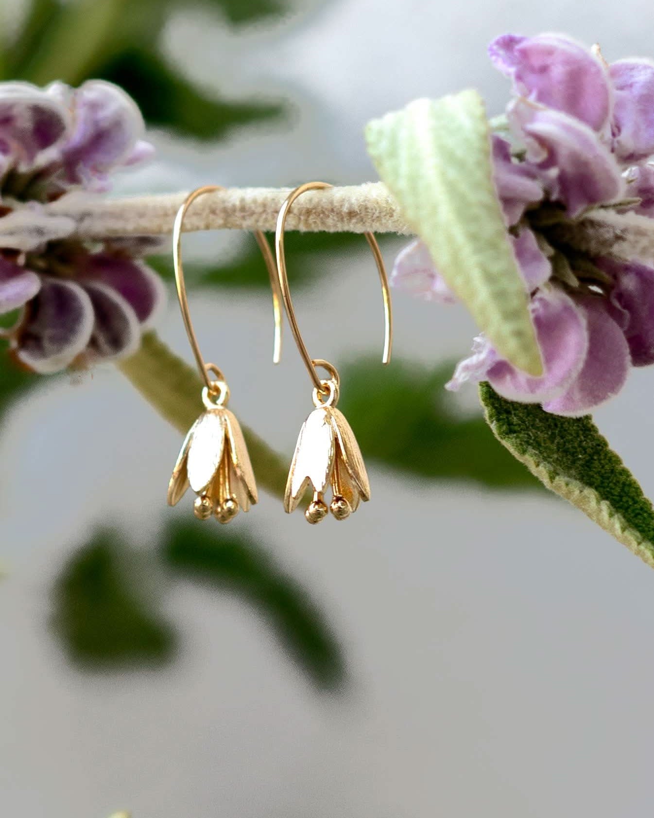 Gold flower-shaped earrings hanging on a branch with purple flowers.