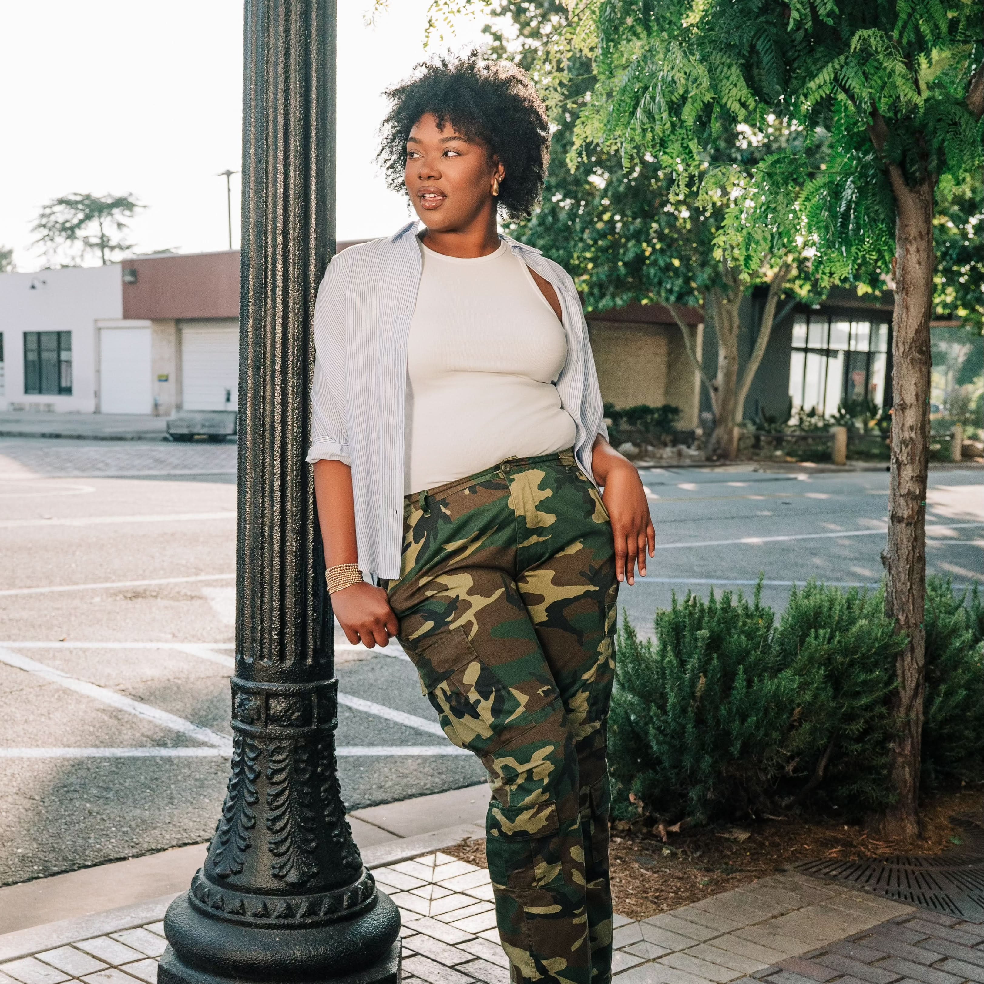 Woman wearing a white ribbed high neck tank top with a white and blue vertical button down with camo pants and black shoes.
